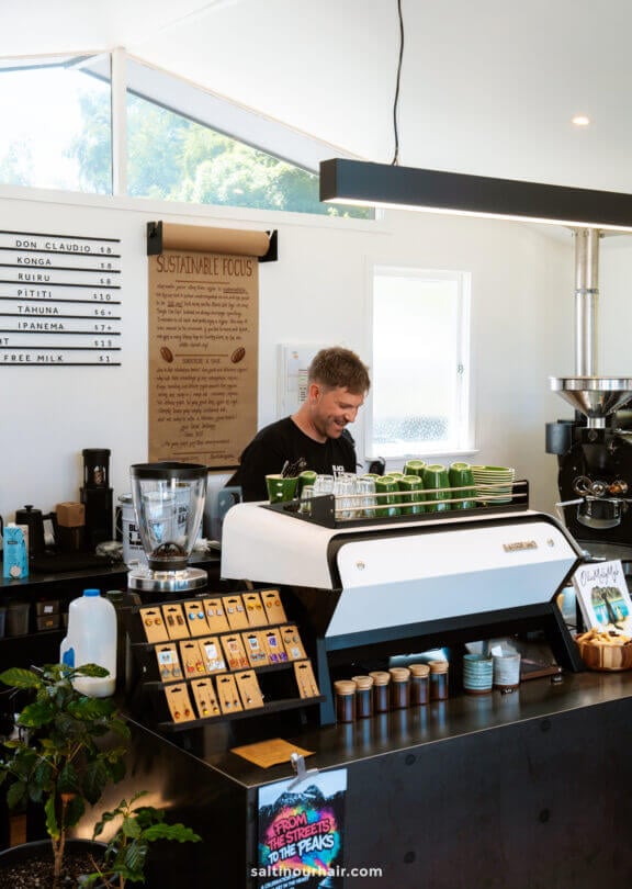 A barista stands behind a coffee counter preparing drinks, with coffee beans, a grinder, and a &ldquo;Sustainable Focus&rdquo; sign visible&mdash;a perfect stop for coffee lovers exploring things to do in Queenstown&rsquo;s modern caf&eacute; scene.