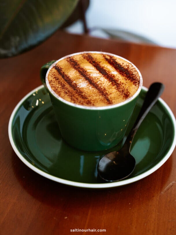 A green cup and saucer with a cappuccino topped with cocoa powder stripes, accompanied by a black spoon on a wooden table&mdash;perfect for relaxing after exploring things to do in Queenstown.