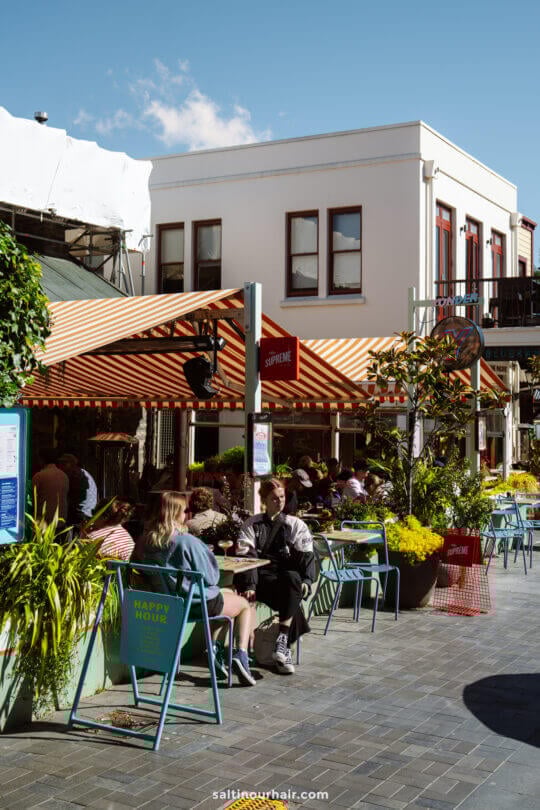 People are sitting at outdoor tables at restaurant yonder, under a striped awning at a lively caf&eacute;, surrounded by plants on a sunny day&mdash;one of the charming things to do in Queenstown, new zealand