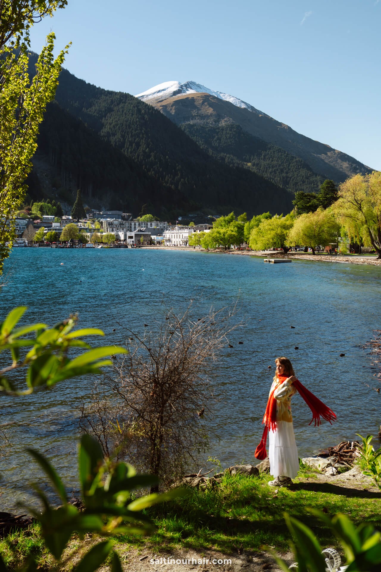 A woman in a white outfit and red scarf stands by a lake with queenstown and snow-capped mountains in the background under a clear sky in new zealand