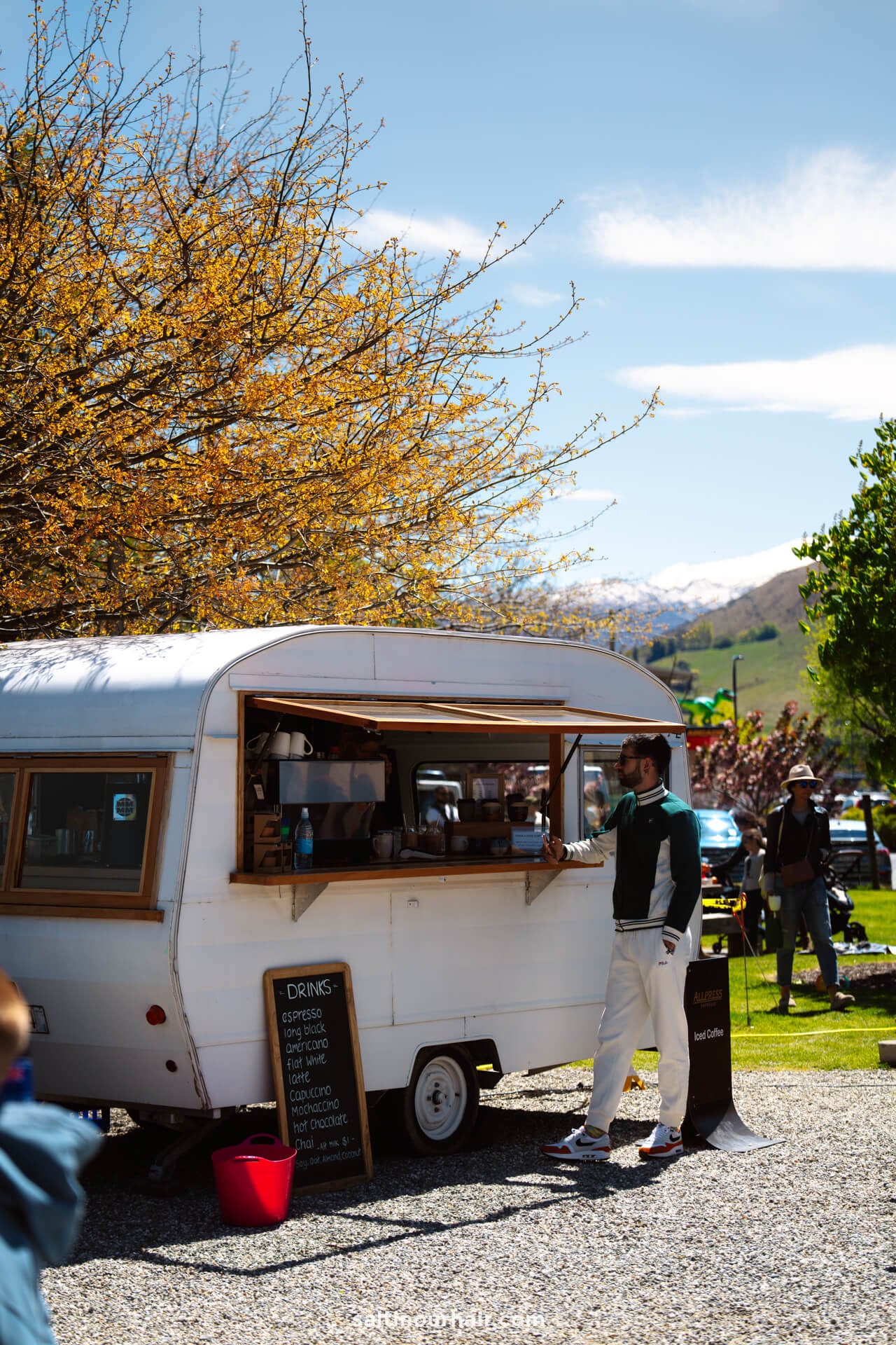 A person stands outside a white food truck serving coffee at Remarkables market, Queenstown, New Zealand with a menu board displayed and trees and mountains in the background