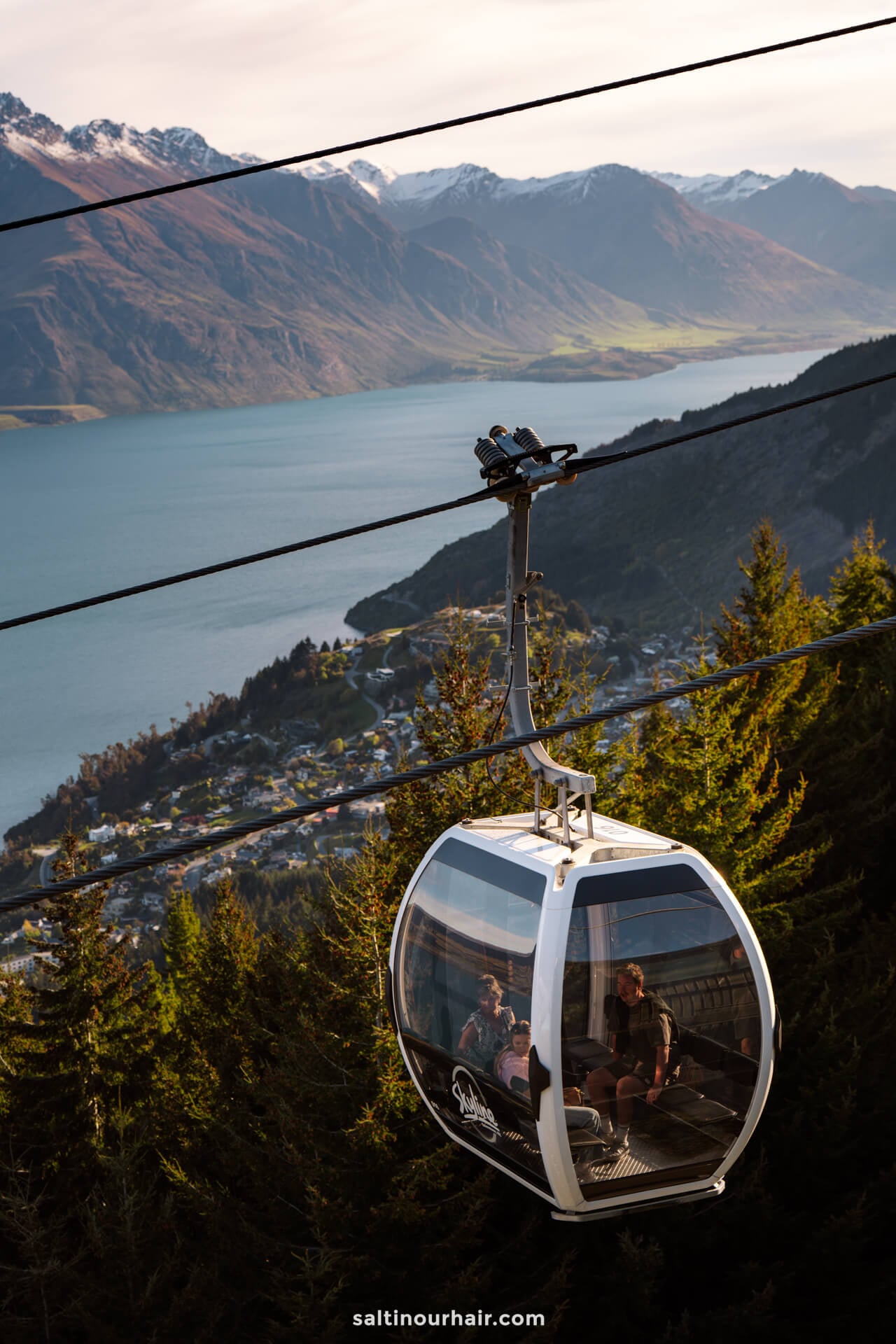 Skyline gondola cable car carrying passengers rises above forested hills, with a lake, a town, and mountains visible in the background&mdash;one of the must-try things to do in Queenstown.