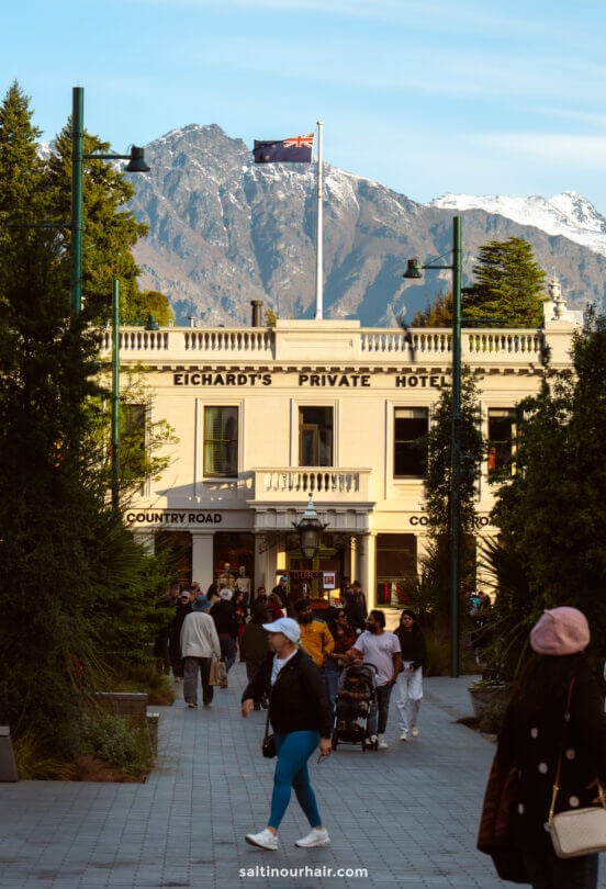 People walk along a tree-lined path toward Eichardts Private Hotel, with mountains and a New Zealand flag in the background&mdash;capturing just one of the many things to do in Queenstown.
