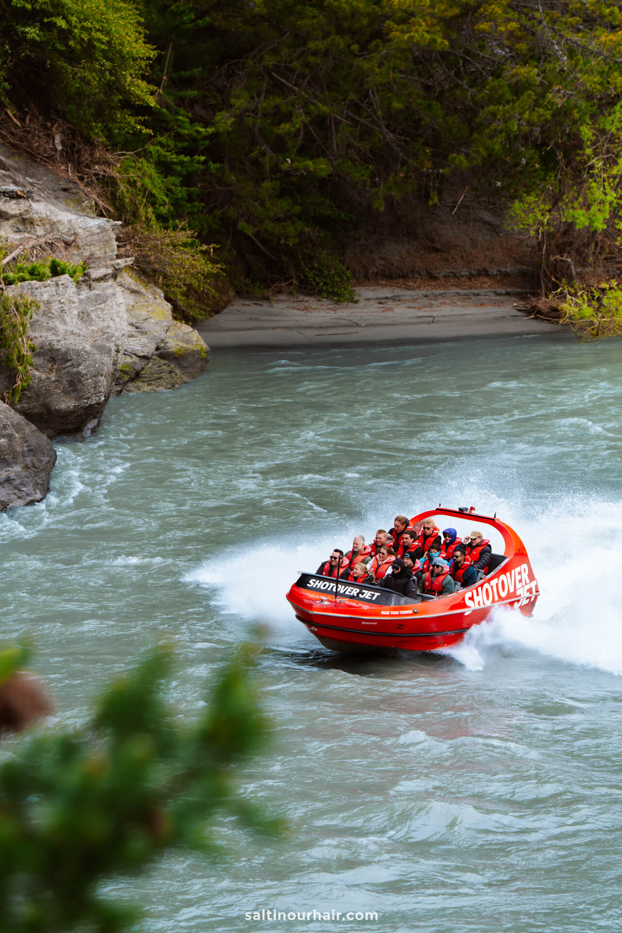 A red jet boat labeled Shotover Jet speeds down a river, carrying passengers in life jackets near rocky, tree-lined banks&mdash;one of the most thrilling things to do in Queenstown, new zealand