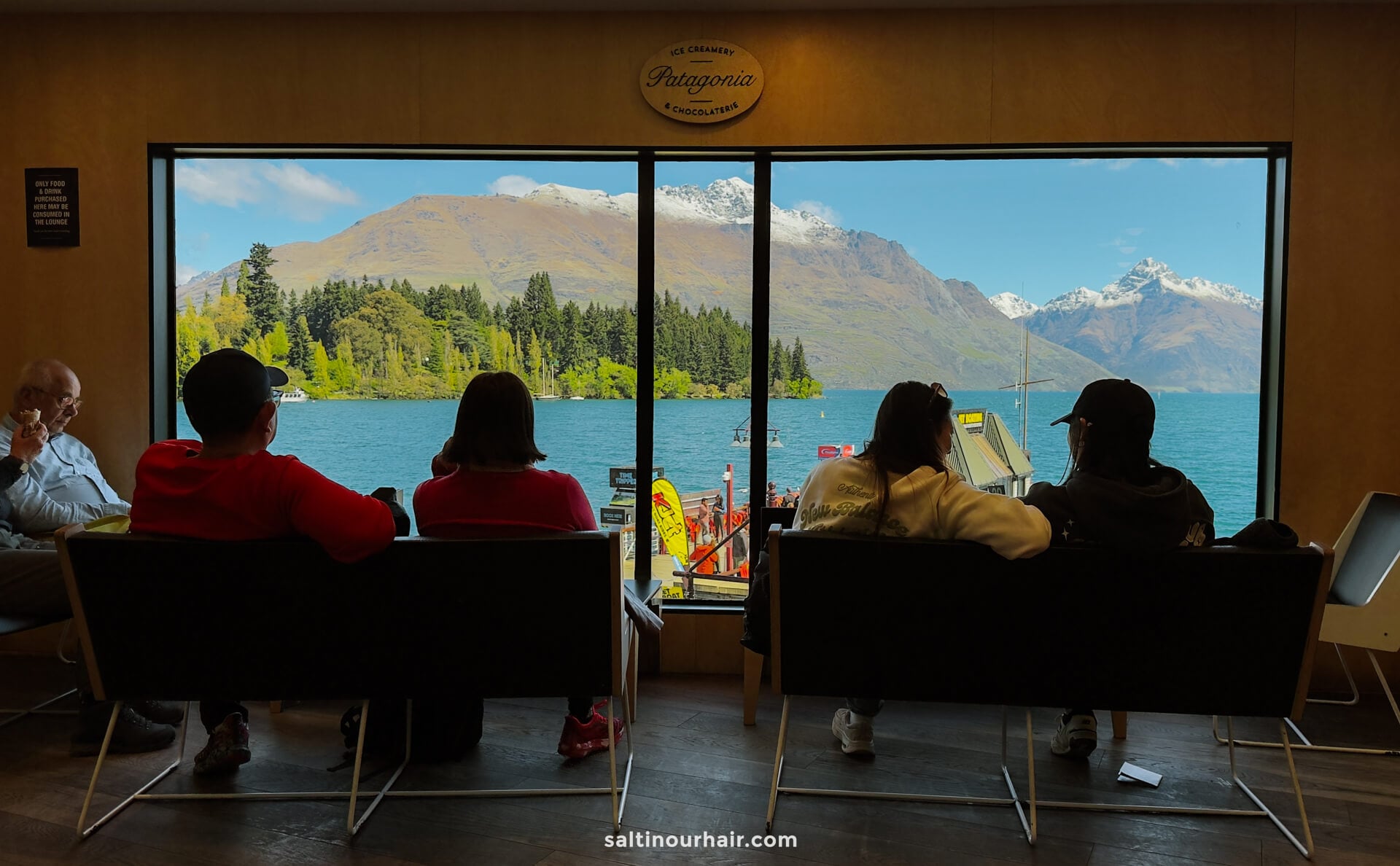 Four people sit indoors on chairs facing large windows at Patagonia Chocolates with a view of a lake, mountains, and a docked boat under a clear sky&mdash;capturing one of the best things to do in Queenstown.