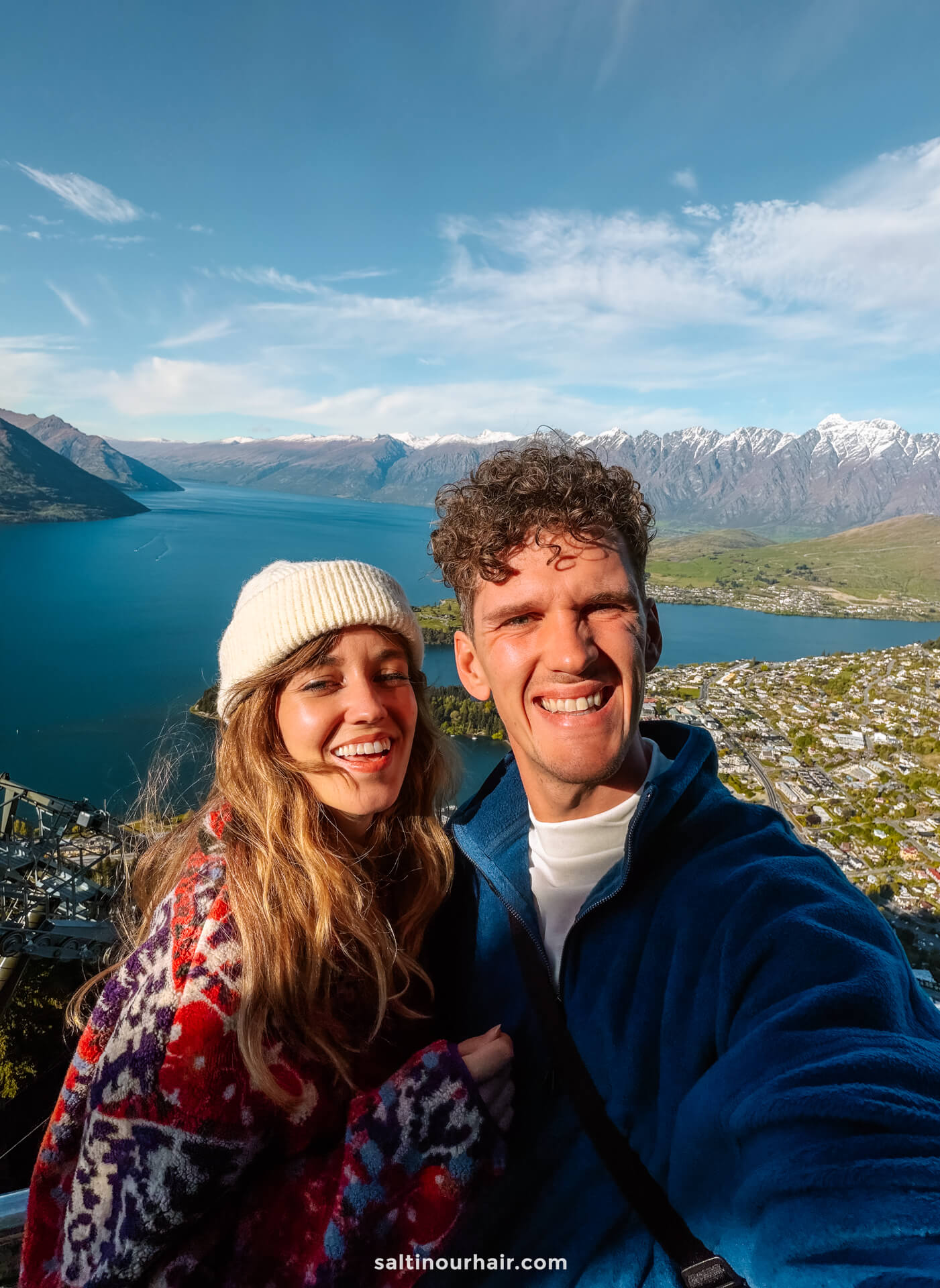 A smiling man and woman take a selfie on a hilltop with a lake, town, and snow-capped mountains in the background under a blue sky, capturing one of the best things to do in Queenstown.