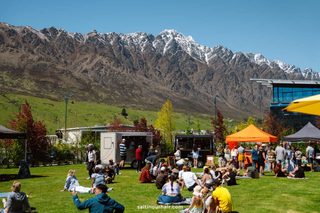 People sit and gather on a grassy field near food trucks and tents at Remarkables Market, with mountains and clear blue sky in the background&mdash;a perfect spot to enjoy some of the best things to do in Queenstown, new zealand