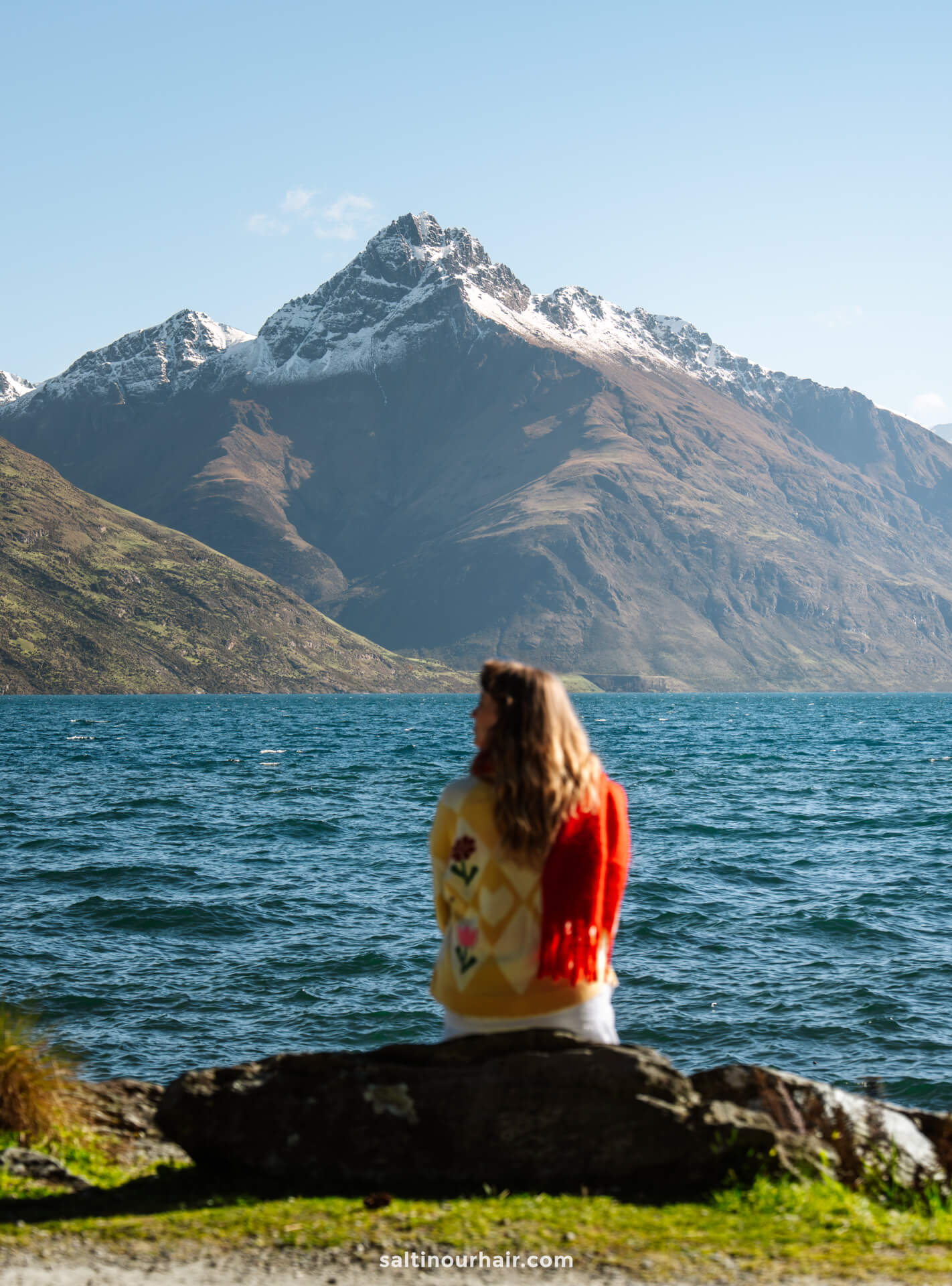 A woman sits on a rock by a lake, facing snow-capped mountains under a clear sky&mdash;one of the unforgettable things to do in Queenstown, new zealand.