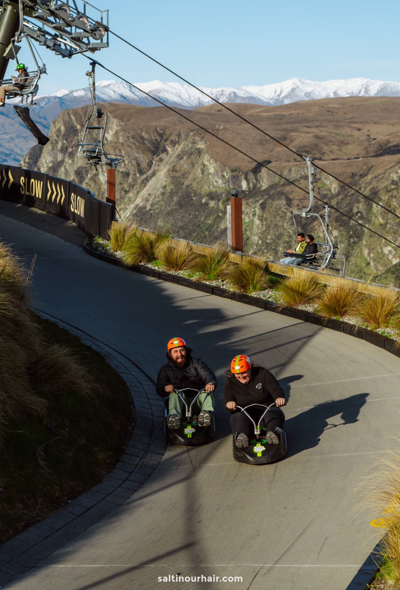 Two people wearing helmets ride luges down a paved mountain track, while another enjoys a ski lift in the background under clear skies&mdash;a thrilling example of things to do in Queenstown, new zealand