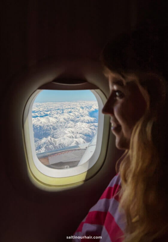 A person sits by an airplane window, looking out at a clear view of snow-capped mountains of new zealand below, dreaming of all the exciting things to do in Queenstown.