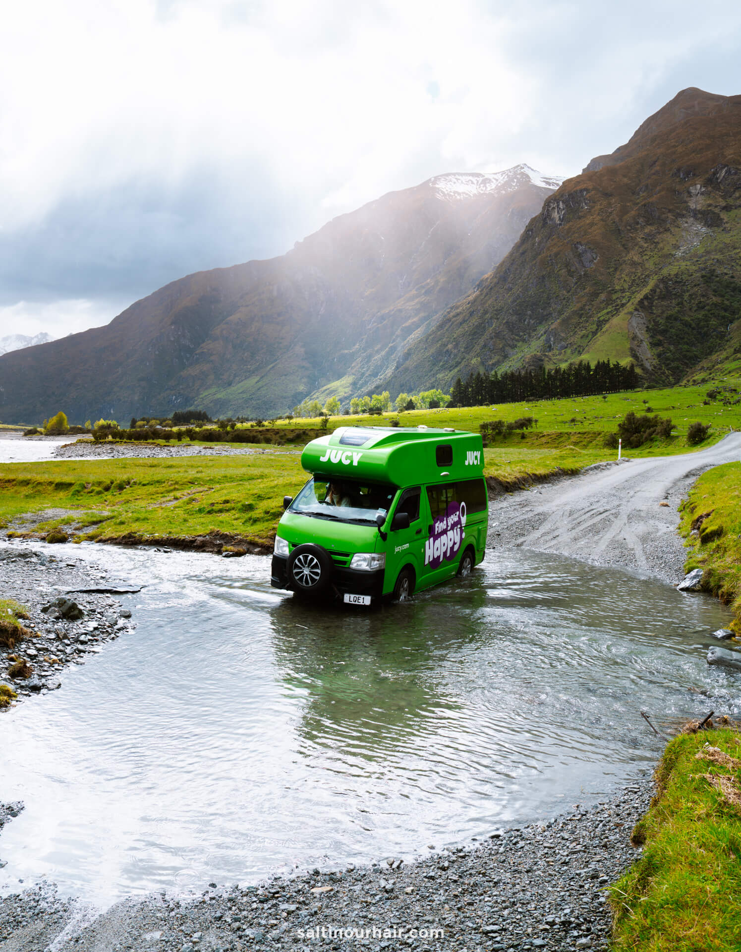 A bright green camper van drives through a shallow stream on the gravel Rob Roy Track in New Zealand,, surrounded by green hills and mountains under a cloudy sky.
