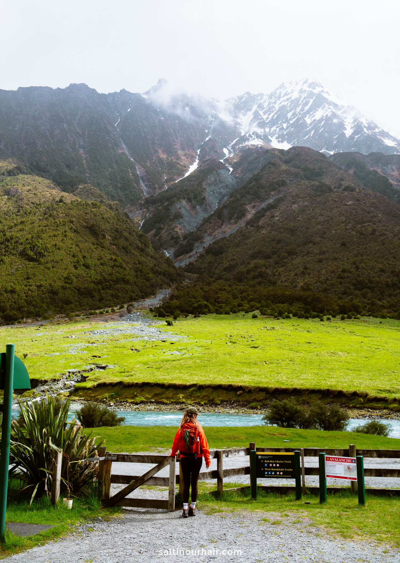 A person with a backpack stands at a gate overlooking the grassy expanse of the Rob Roy Track, with a river and snow-capped mountains beneath an overcast sky.