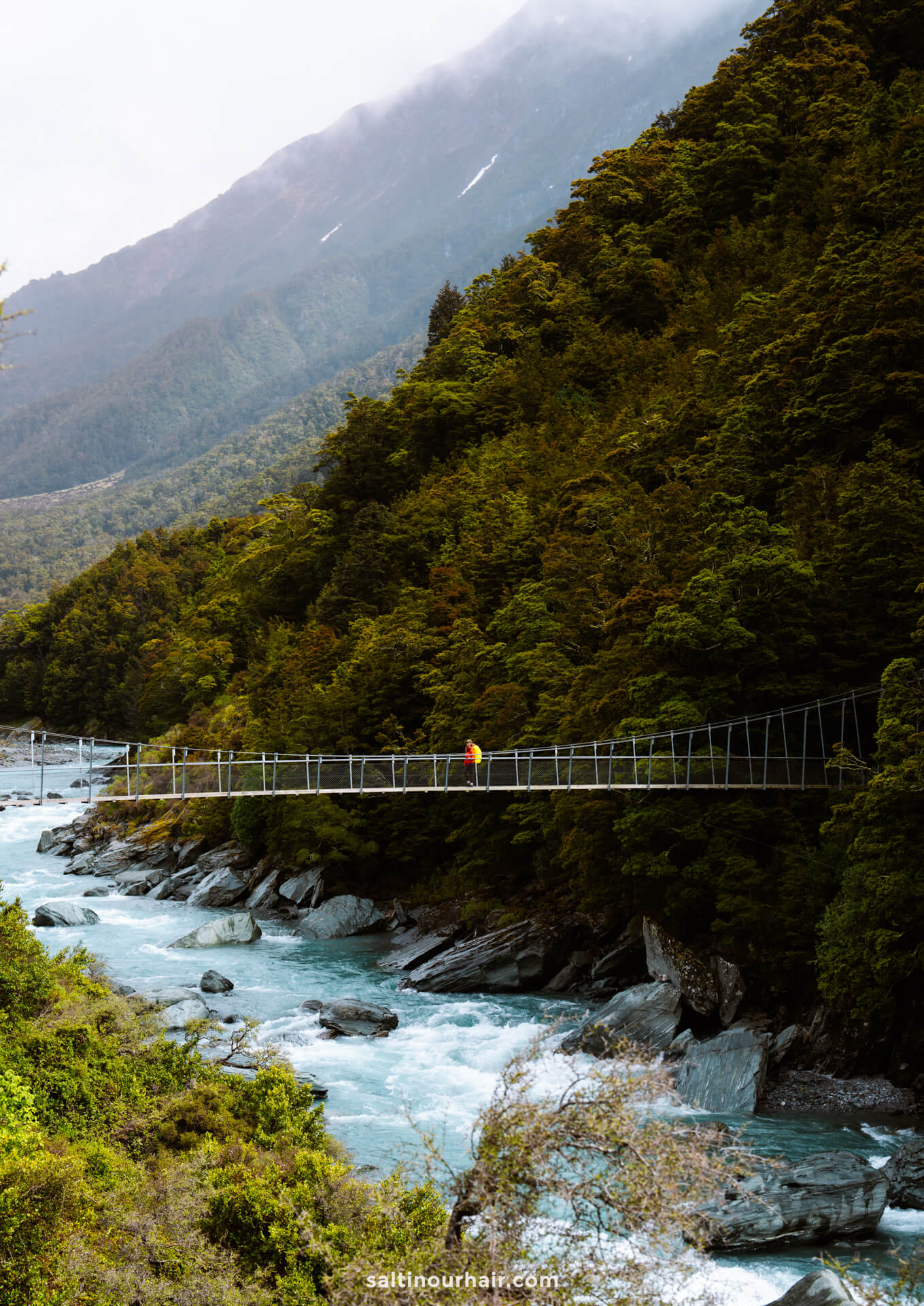 A person in a yellow jacket crosses a suspension bridge on the Rob Roy Track over a blue river, surrounded by dense forest and mountains.