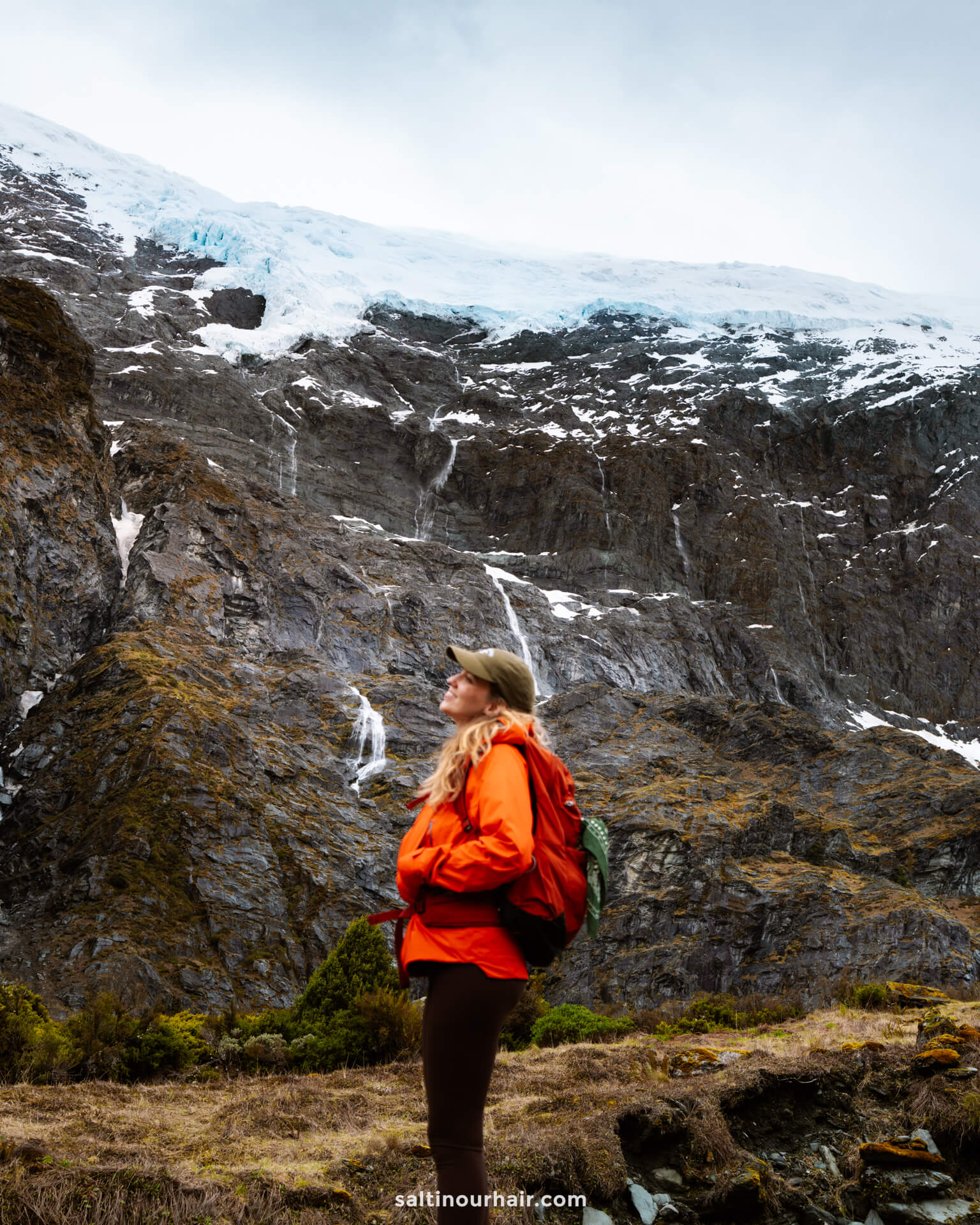 A person in an orange jacket and backpack stands on grassy ground, looking up at a rocky mountain with patches of snow and ice under a cloudy sky along the scenic Rob Roy Track in New Zealand