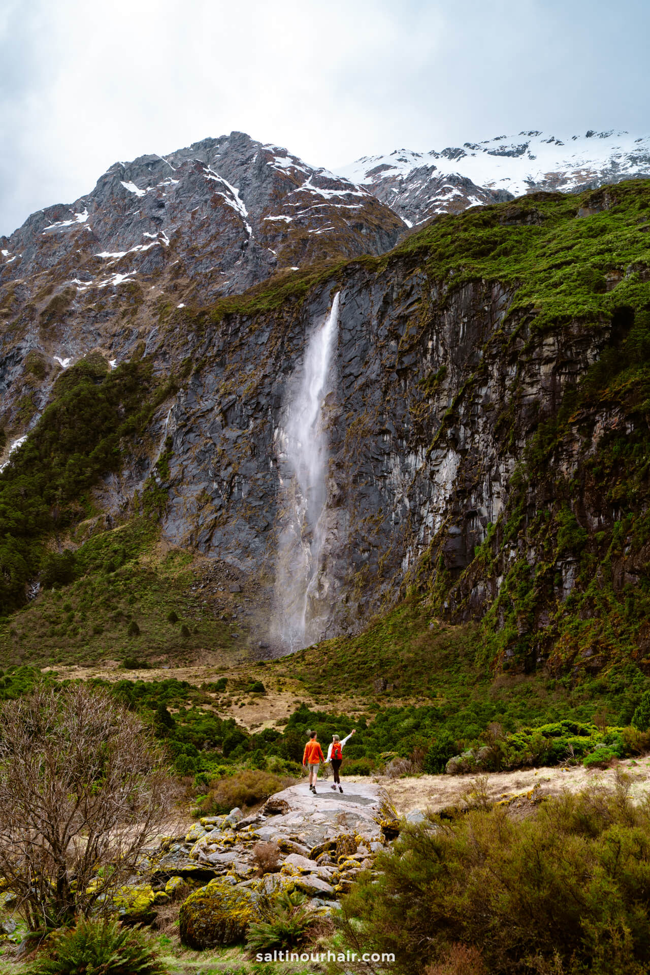 Two people in red jackets walk along the Rob Roy Track in New Zealand, heading toward a tall waterfall cascading down a rocky, green mountainside topped with snow.