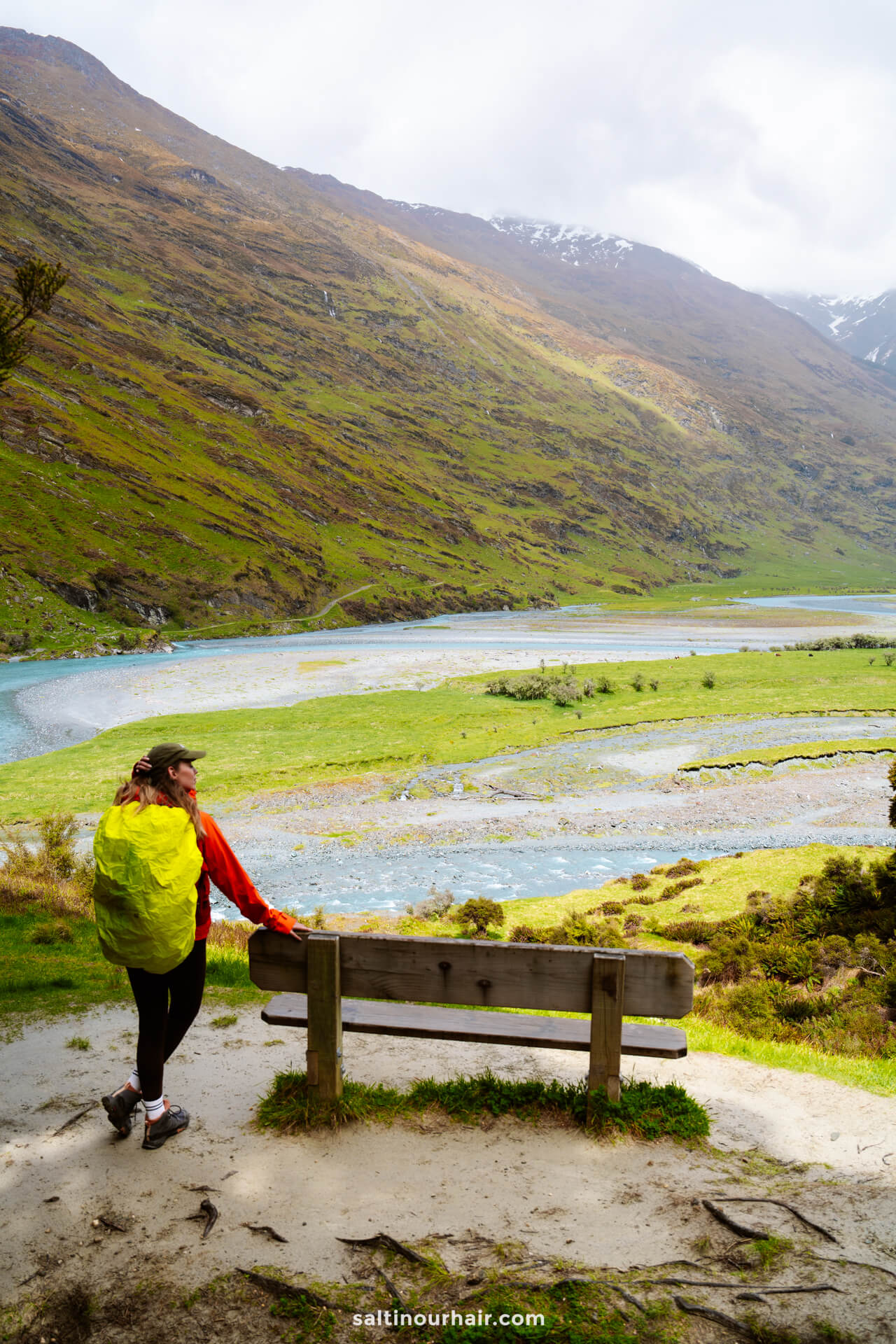 A person with a yellow backpack stands by a wooden bench overlooking the winding river and green valley of the Rob Roy Track in New Zealand, surrounded by mountains.