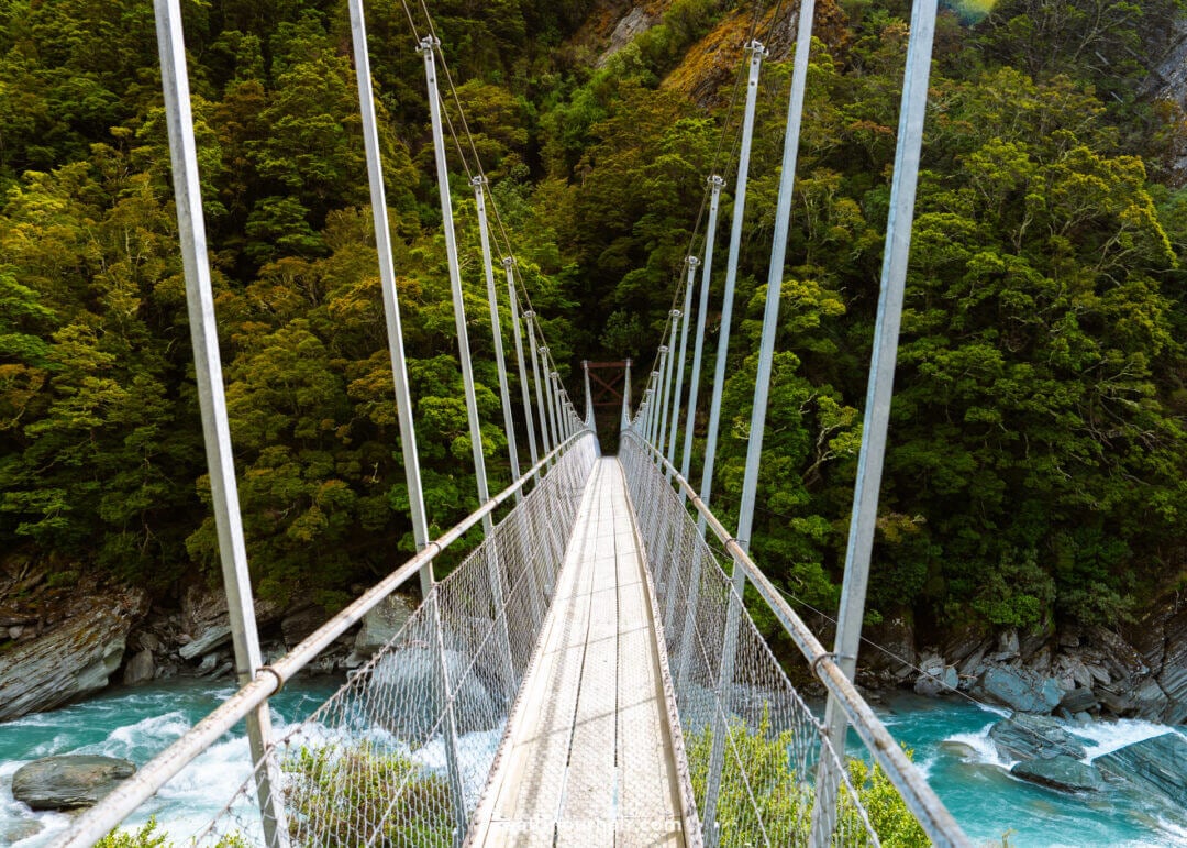 A suspension footbridge crosses over a river with rocks below, surrounded by dense green forest and steep hillsides along the scenic Rob Roy Track in New Zealand,