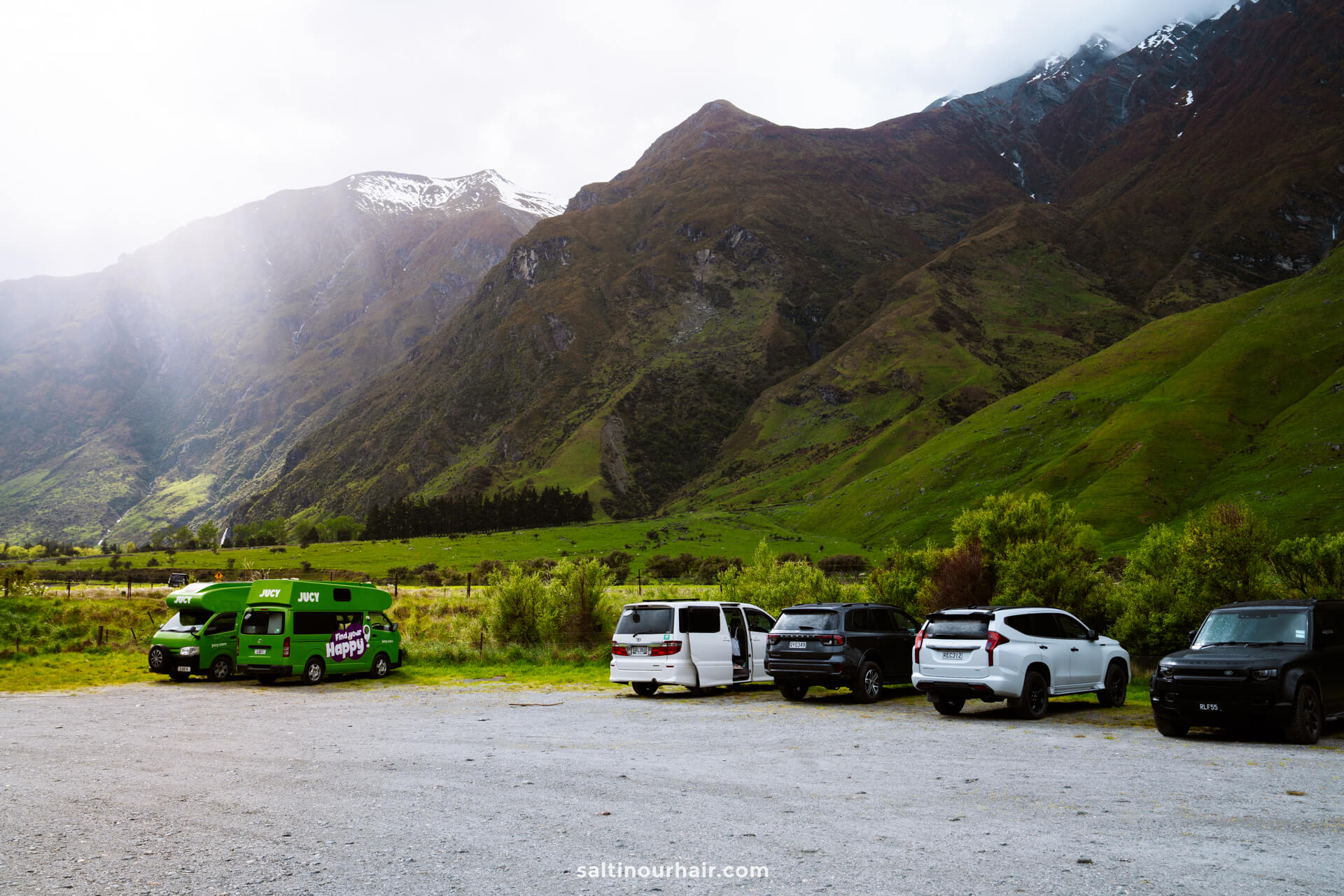 A row of parked vehicles, including a green camper van, sits on a gravel lot near the start of the Rob Roy Track, framed by green mountains and snow-capped peaks under a partly cloudy sky.