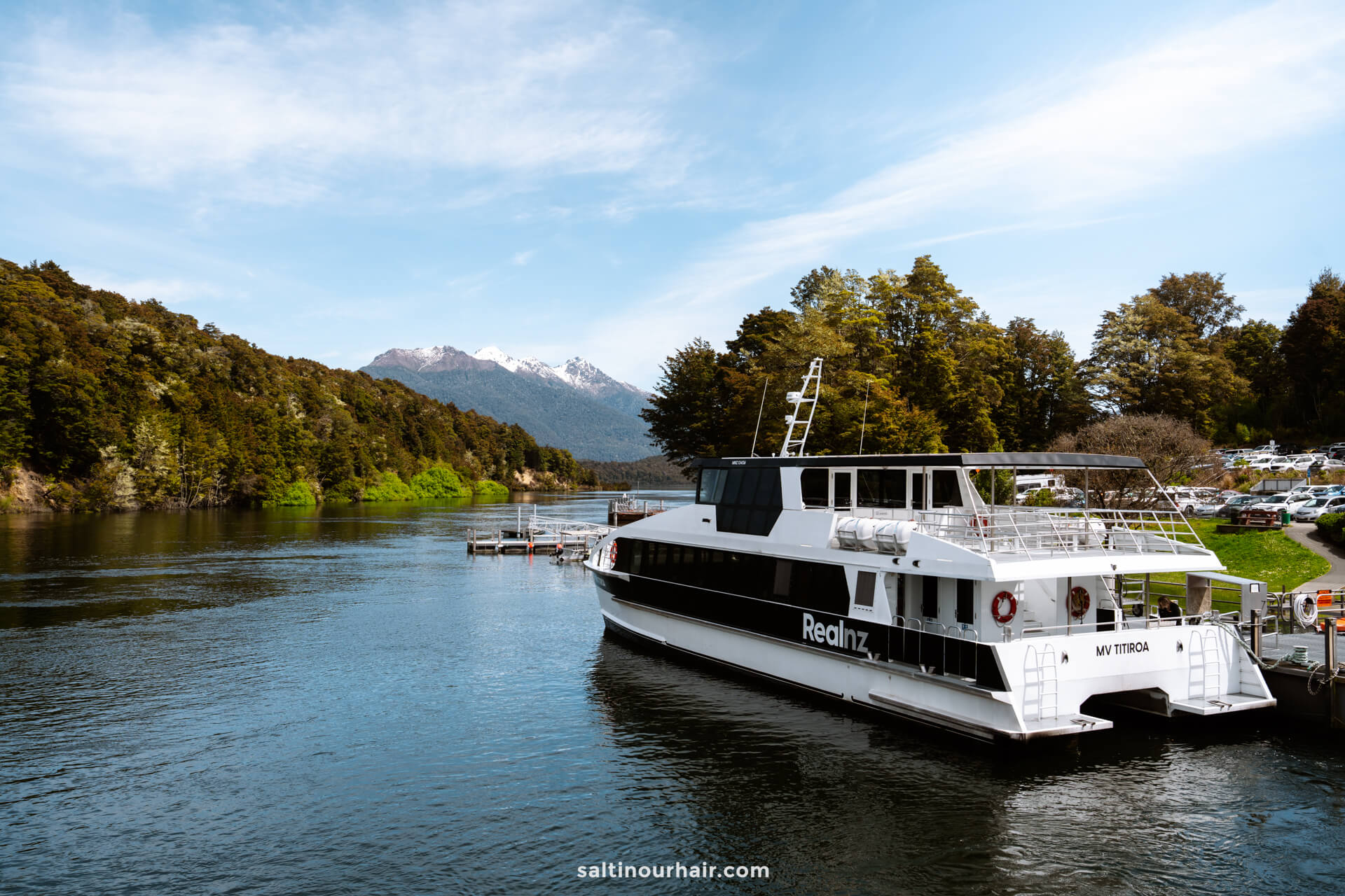 A white tour boat named RealNZ is docked on the calm waters of lake manapouri surrounded by trees, with mountains and a partly cloudy sky in the background.