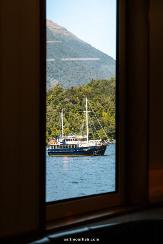 A sailboat floats on a lake near a forested mountain, seen through a window frame&mdash;evoking the tranquility of an overnight cruise in Doubtful Sound, New Zealand.