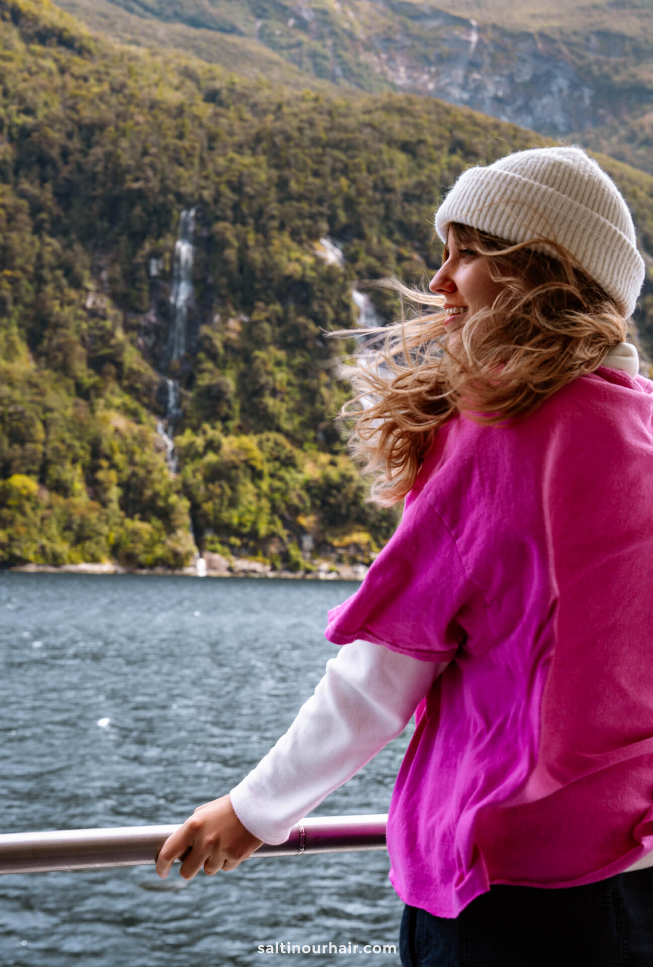 A person wearing a pink shirt and white beanie stands on a boat during an overnight cruise in Doubtful Sound, New Zealand, smiling with water and green hills in the background.