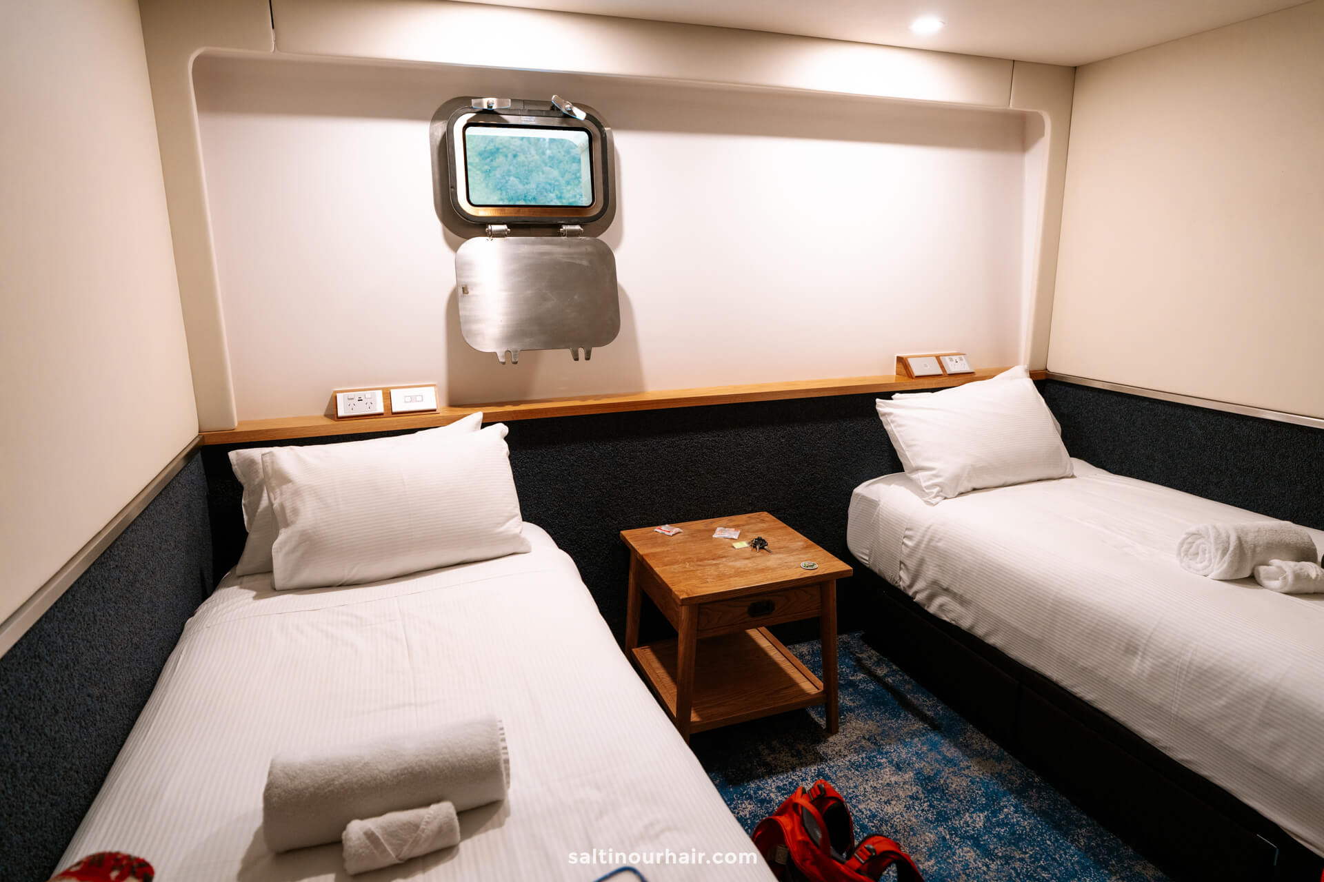 A small cabin room with two single beds, white linens, a wooden nightstand between them, a small window above, and towels placed on each bed&mdash;perfect for an overnight cruise on Doubtful Sound, New Zealand.