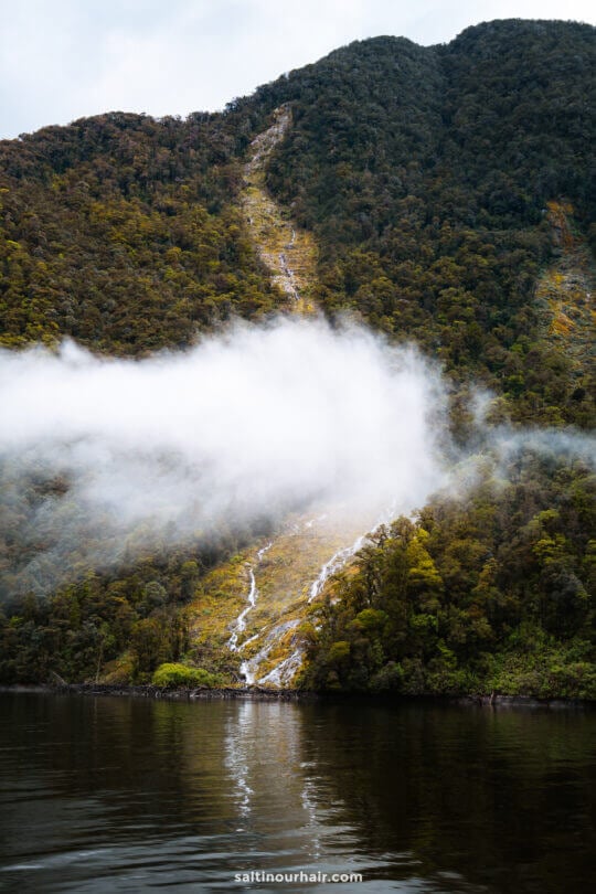 A narrow waterfall flows down a green mountainside partially covered by a low, horizontal cloud, with its base meeting a calm dark lake.