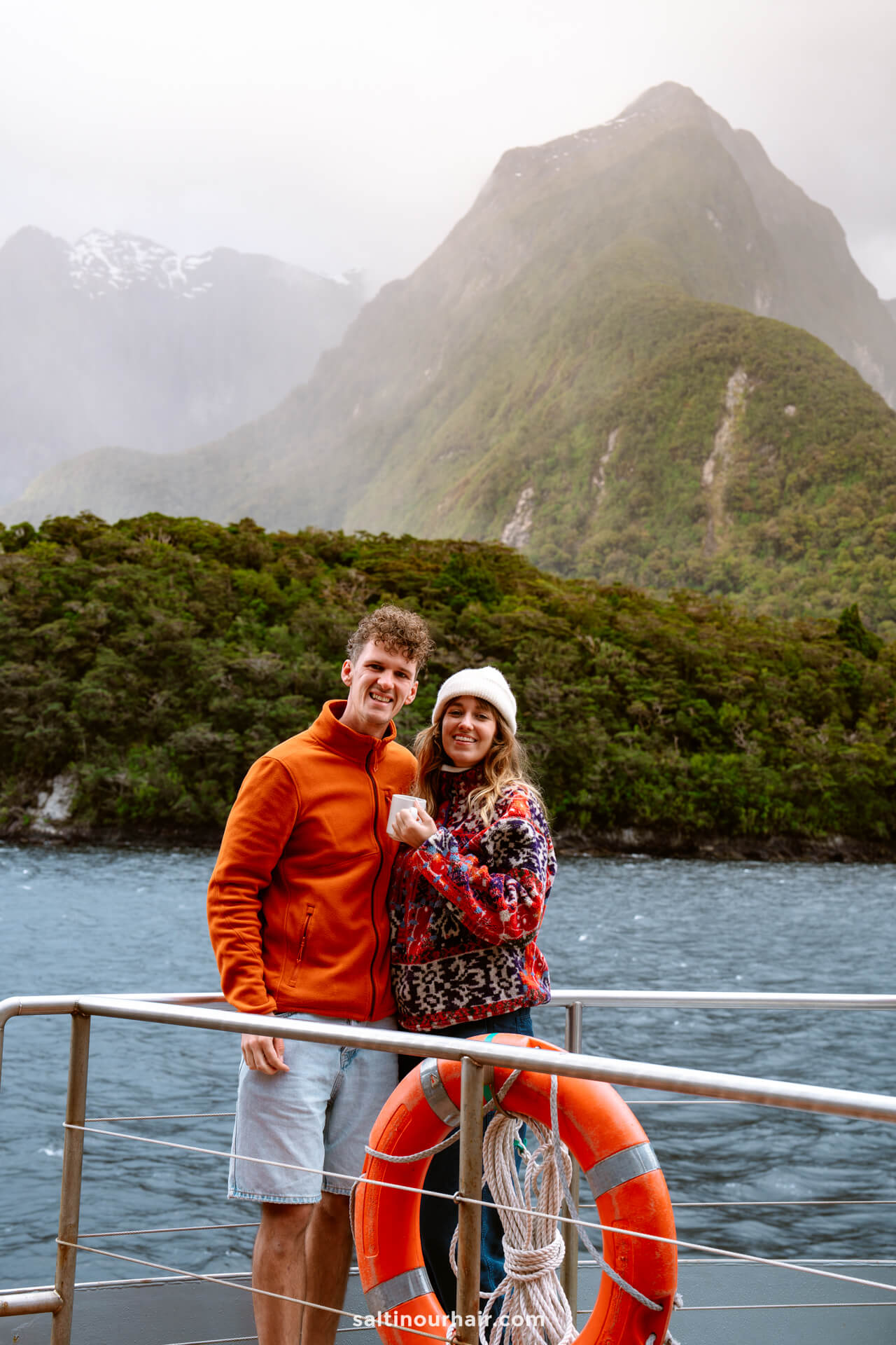 Two people stand on a boat deck of the overnight cruise in doubtful sound in front of a mountainous landscape, smiling and holding hands near an orange life preserver.