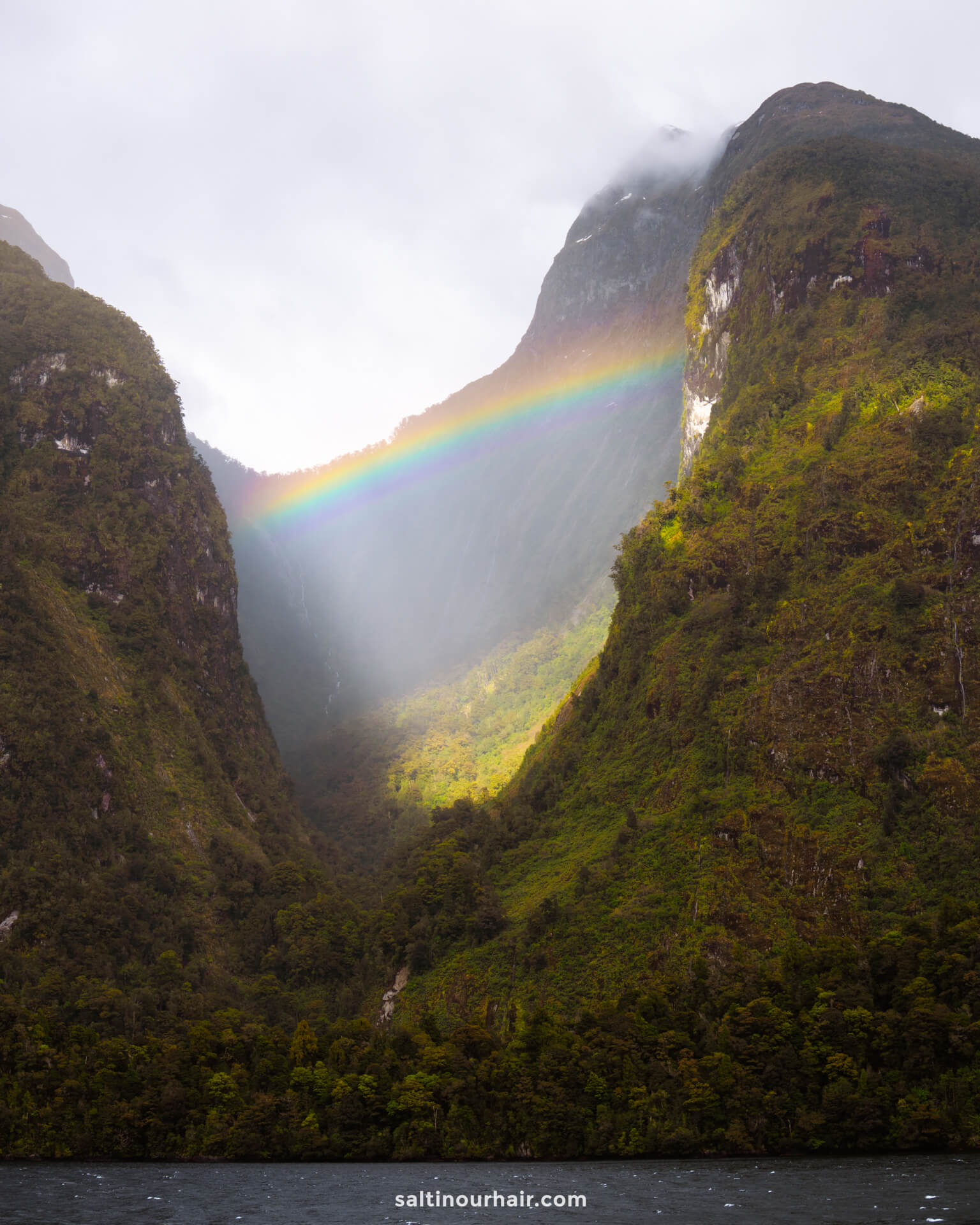 A rainbow appears between two forested mountains under a cloudy sky, with sunlight breaking through and illuminating the scene. an incredible scene during an overnight cruise in doubtful sound new zealand