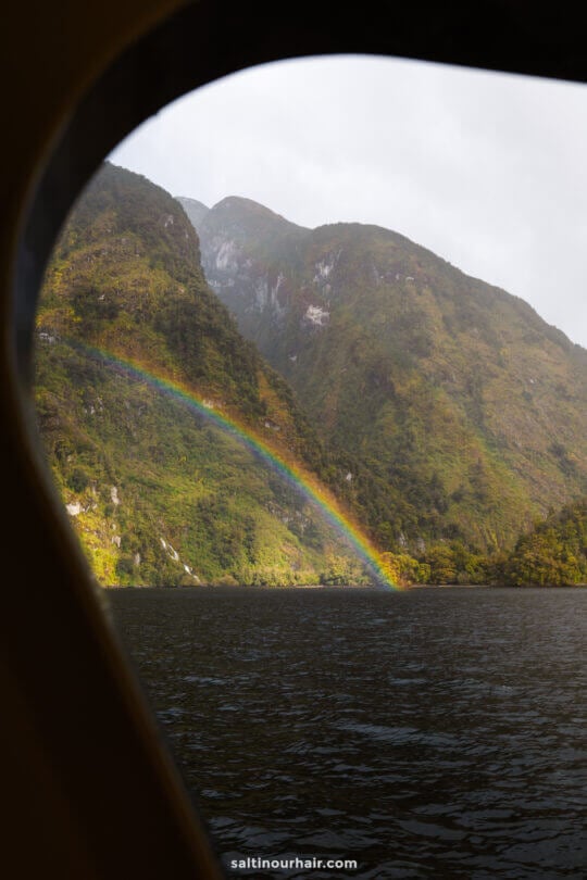 A rainbow arcs over green mountains and a lake, viewed through the window of a boat.