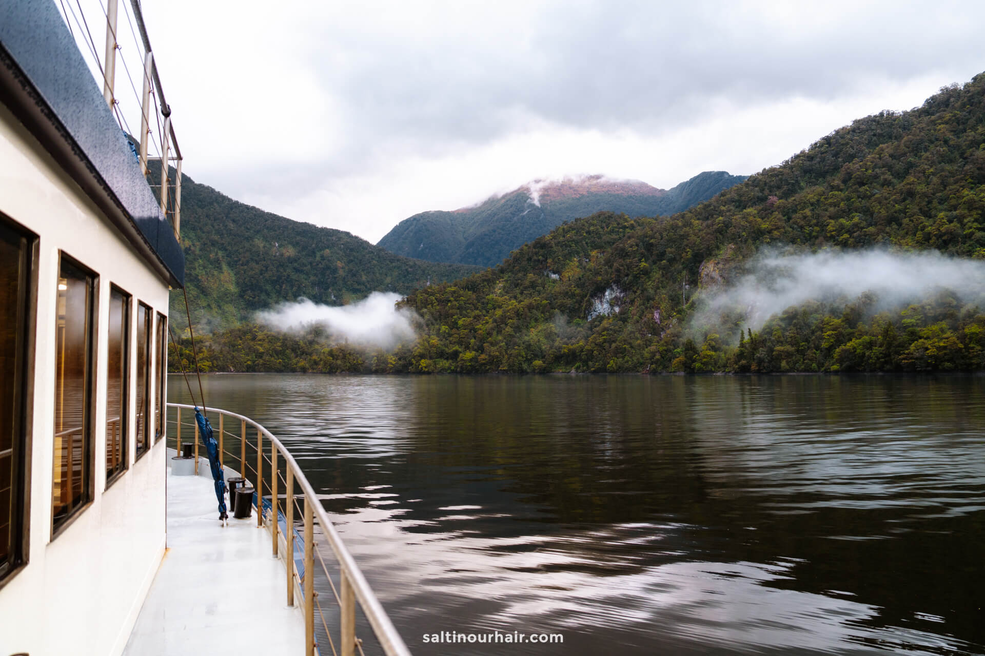 A boat cruises on a calm lakes of doubtful sound, new zealand surrounded by green forested hills and low clouds under an overcast sky.