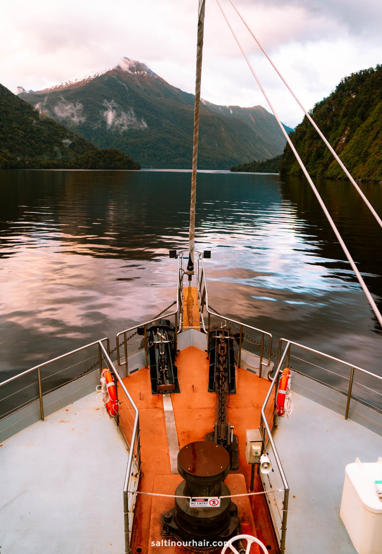 Experience the breathtaking view from a boat&rsquo;s deck on an overnight cruise in Doubtful Sound, New Zealand, with calm waters reflecting forested mountains and a distant snow-capped peak beneath a cloudy sky.