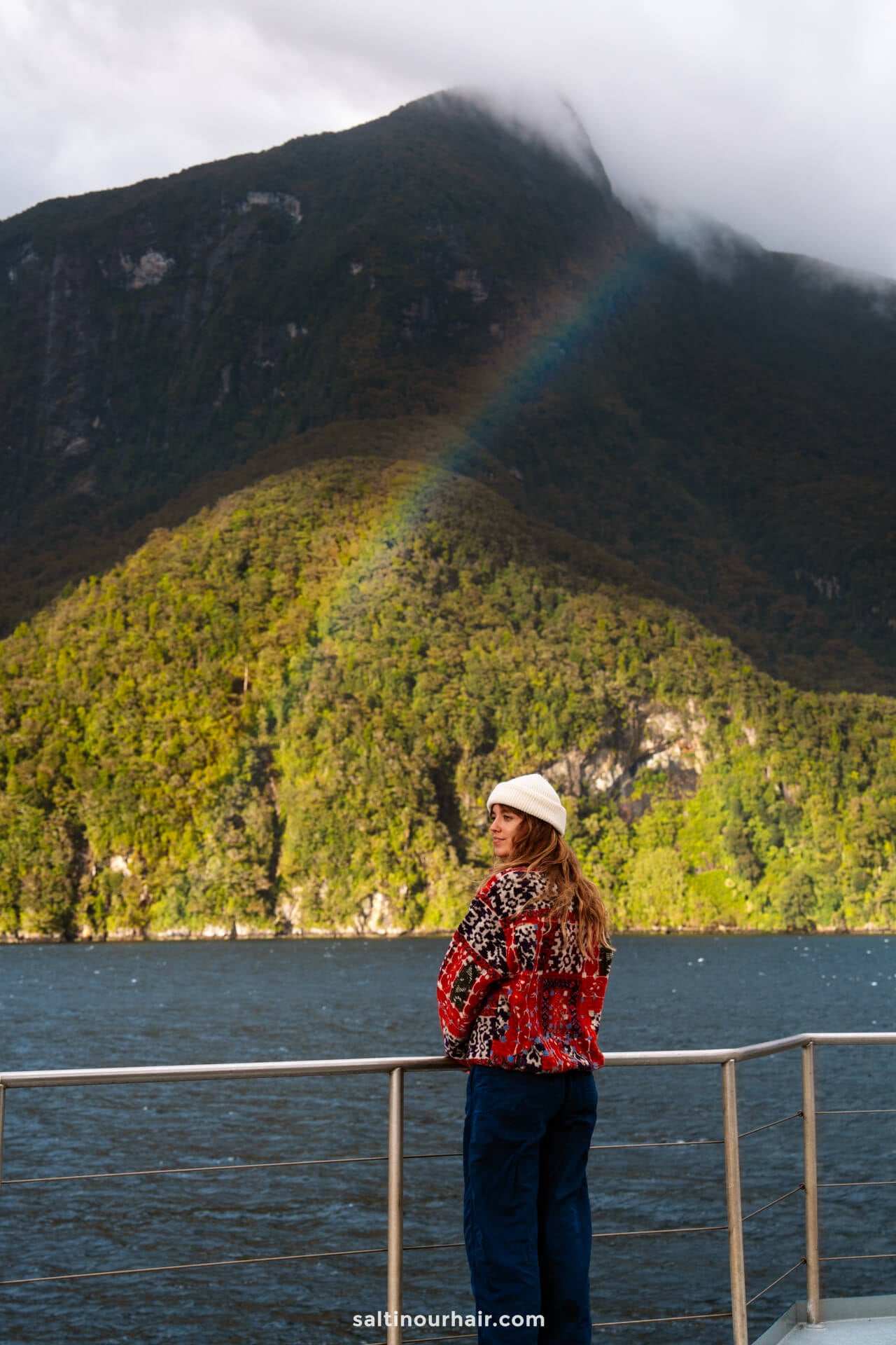A person in a red patterned sweater and white beanie stands on the deck of an overnight cruise in Doubtful Sound, New Zealand, gazing at a green hill with a faint rainbow in the background.