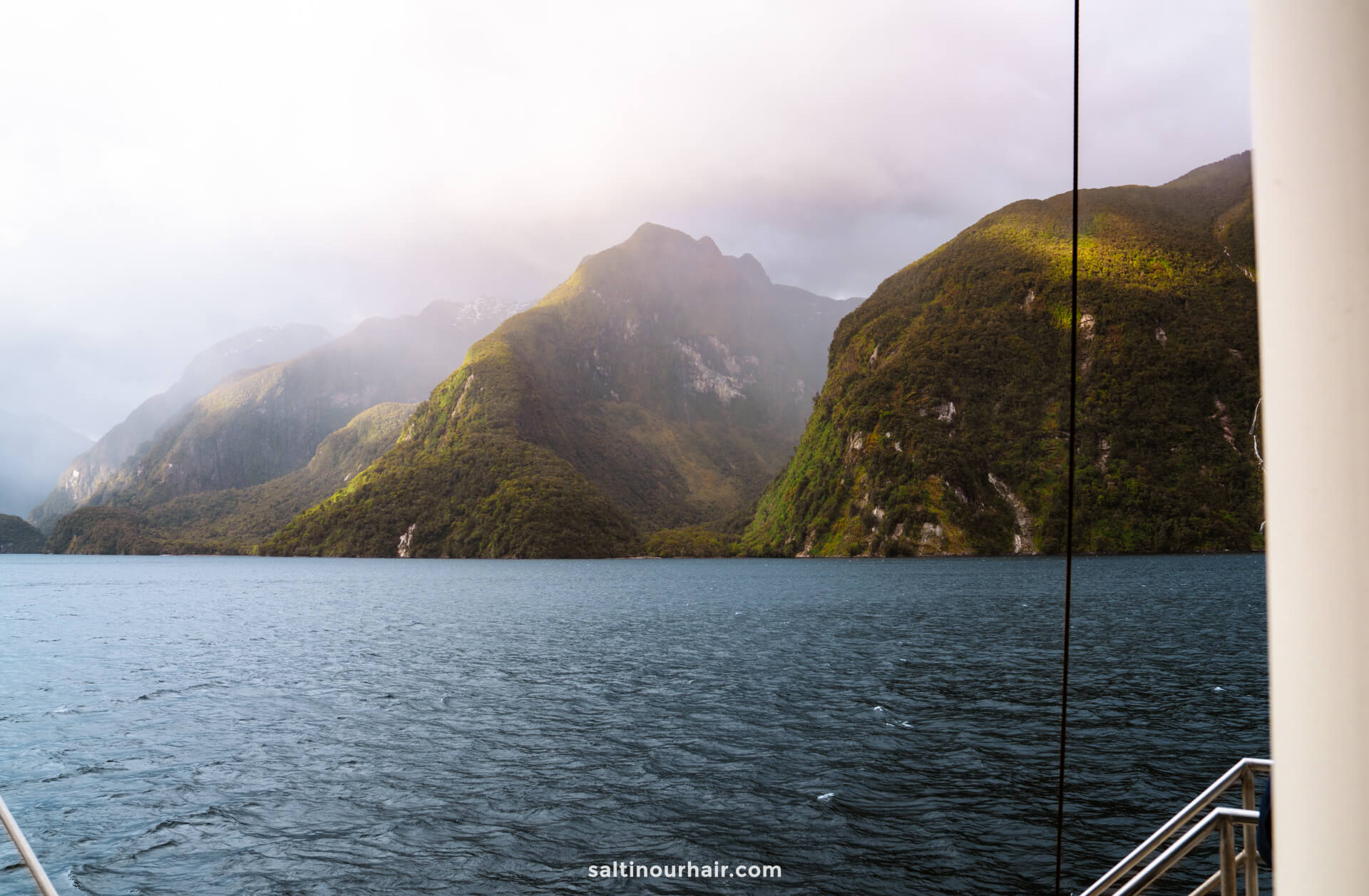 calm water surrounded by green, mountainous cliffs under a cloudy sky, viewed from a boat on an overnight cruise through Doubtful Sound, New Zealand.
