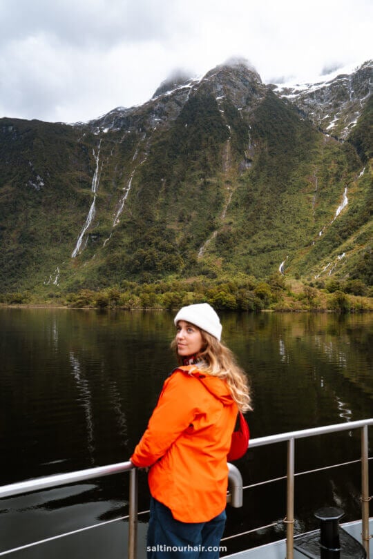 A woman in an orange jacket and white beanie stands on a boat, looking back, with a lake and tall green mountains of doubtful sound in the background.