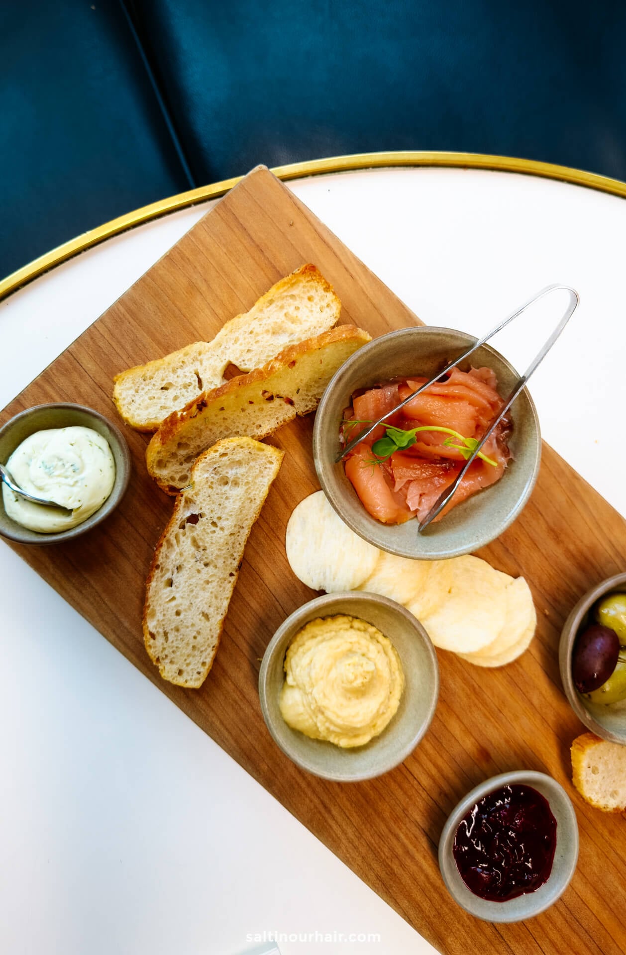 A wooden serving board with slices of bread, chips, olives, smoked salmon with garnish, a dip, cream cheese, and jam on a white table.