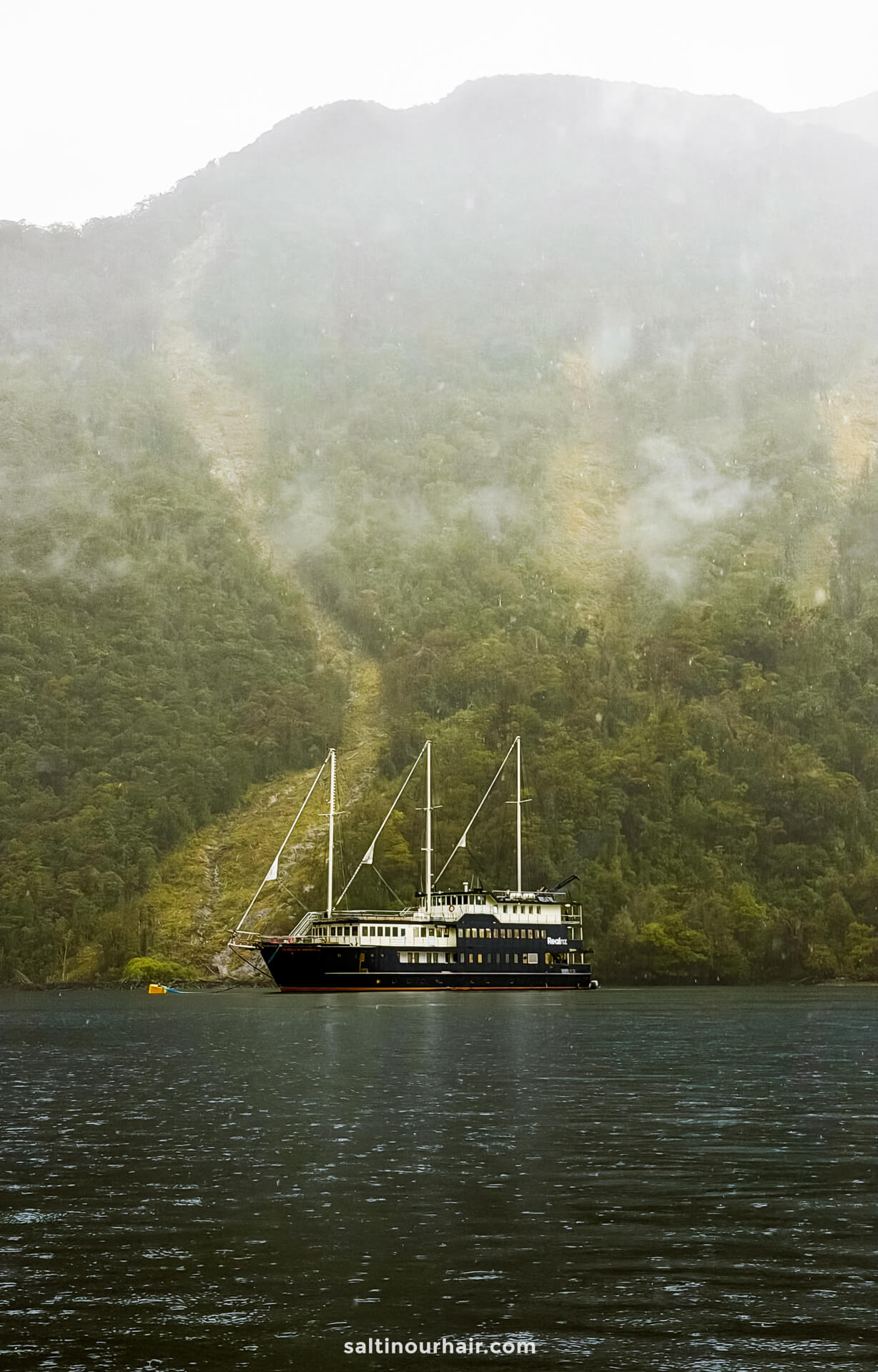 A blue yacht with three masts is anchored on a misty lake, surrounded by dense green forest and steep hillsides&mdash;an ideal scene for an overnight cruise in Doubtful Sound, New Zealand.