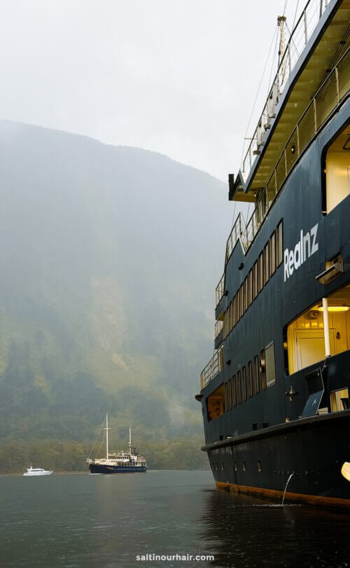 A large ship labeled Realnz is docked by a misty fjord, capturing the serene start of an overnight cruise at Doubtful Sound from Queenstown, New Zealand, with two small boats in the water and stunning mountains in the background.