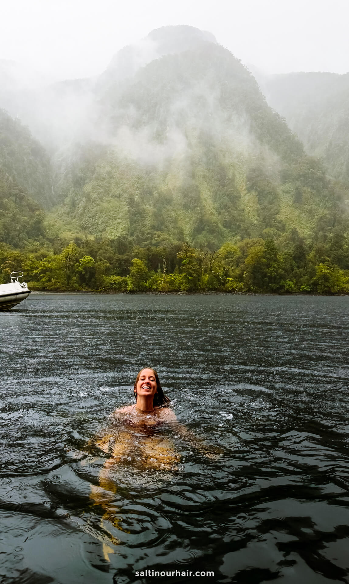 A woman swims in a dark lake of doubtful sound new zealand with forested mountains and mist in the background on a cloudy day.