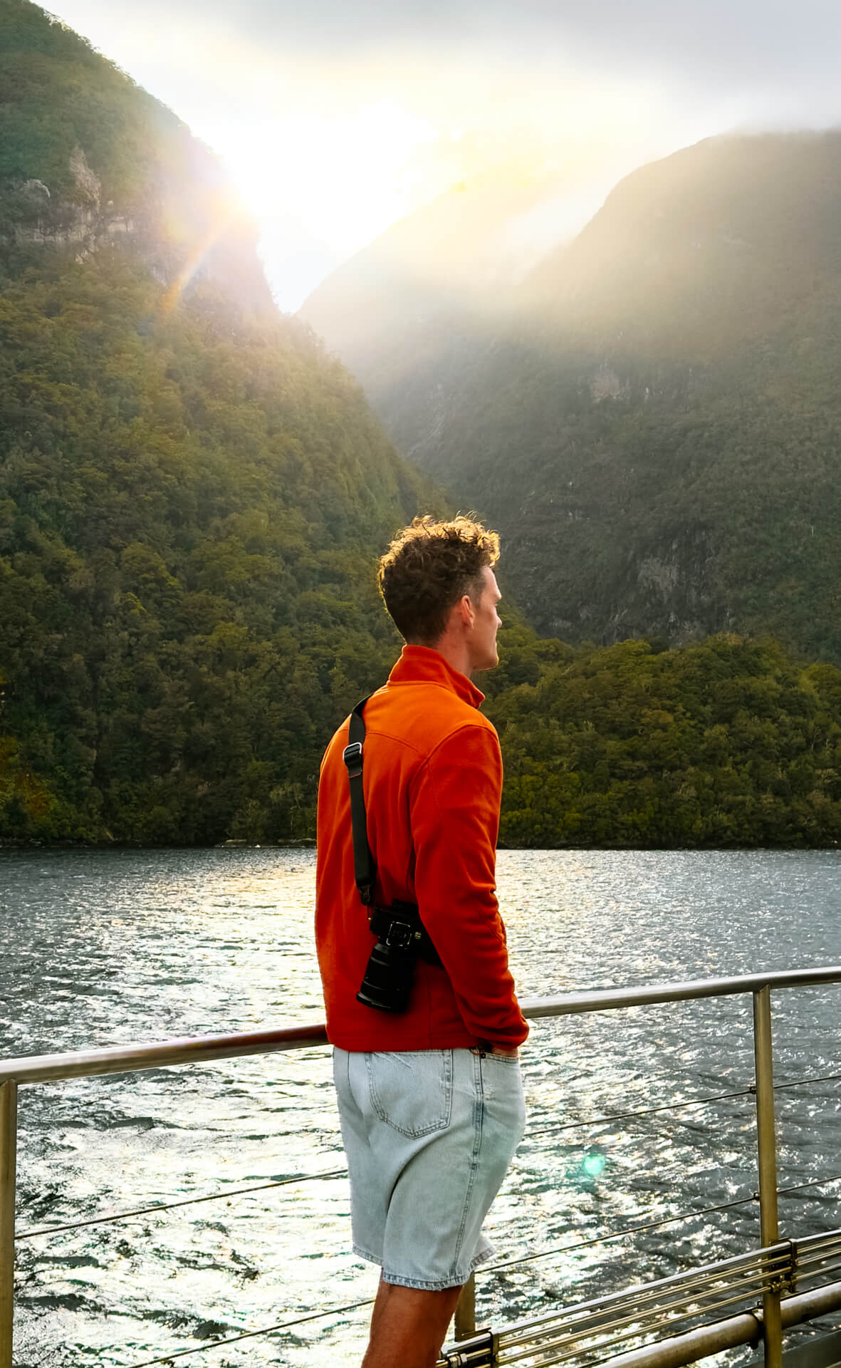 A man in an orange jacket and shorts stands on a boat deck, looking at a lake surrounded by forested mountains under a partly cloudy sky with sunlight streaming through.