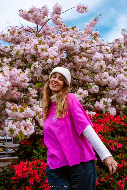 A person in a white beanie and pink shirt stands smiling in front of blooming pink cherry blossoms and red flowers in Te Anau New Zealand