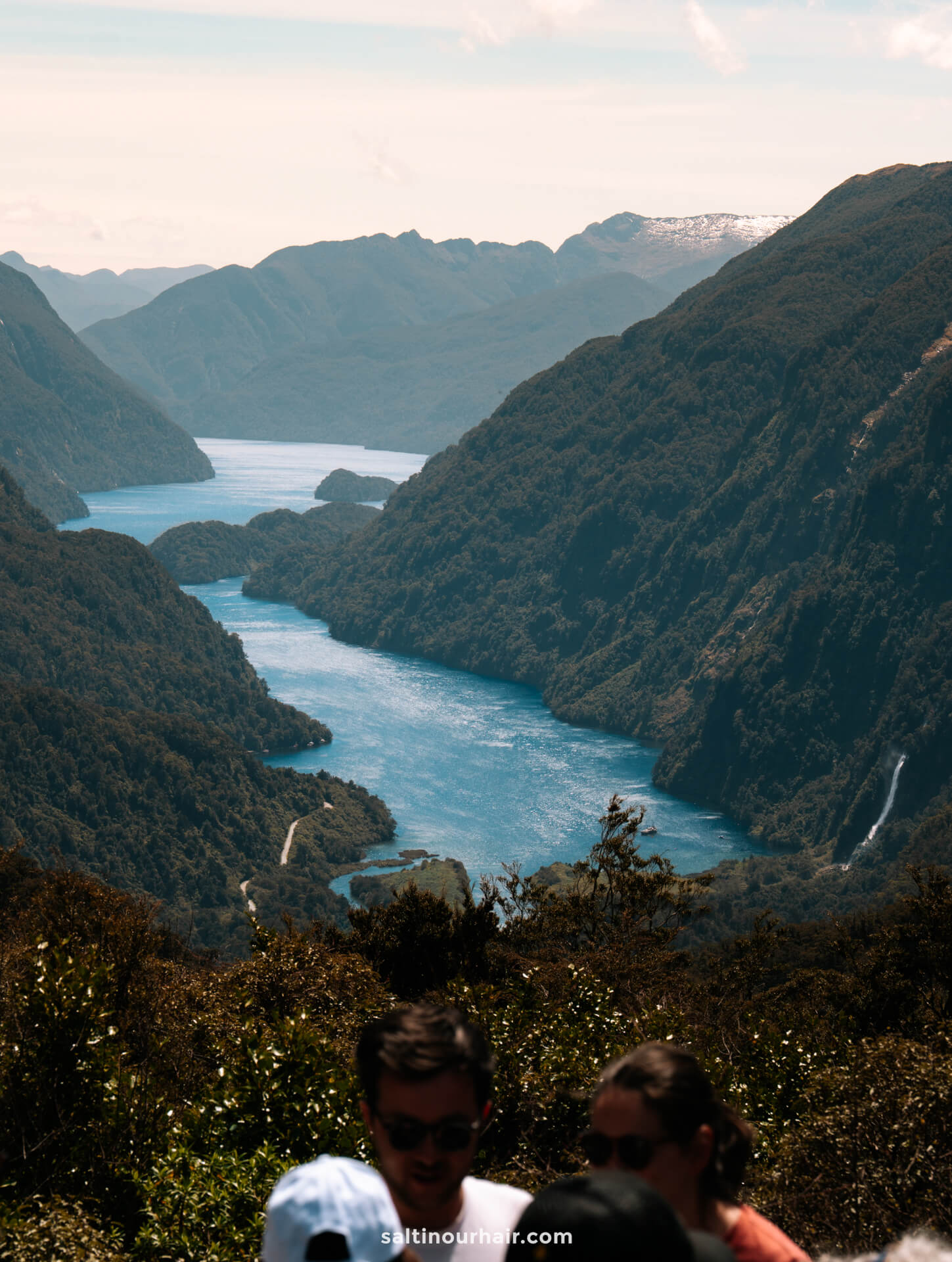 A winding blue lake between forested mountains under a clear sky, reminiscent of Doubtful Sound, with three people in the foreground.
