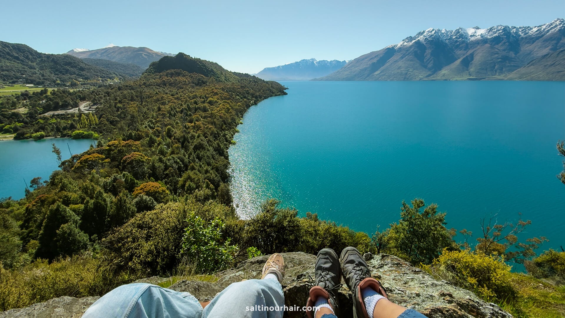Two people sitting on a rocky ledge overlook a large, blue lake surrounded by green hills and distant snow-capped mountains&mdash;a breathtaking hike called Bob