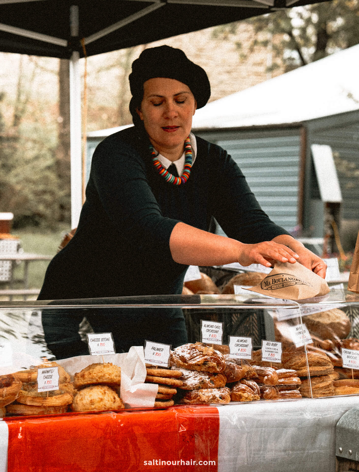 A woman wearing a black beret arranges baked goods at an outdoor market stall in Arrowtown, New Zealand, with various pastries and breads displayed in front of her.
