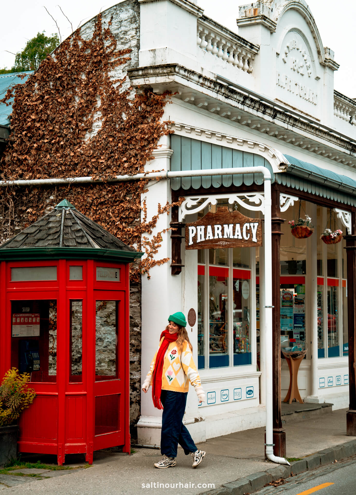 A person walks past a vintage pharmacy with a PHARMACY sign and a red phone booth on the sidewalk in arrow town&mdash;just one of the charming sights among the unique things to do in Queenstown, new zealand
