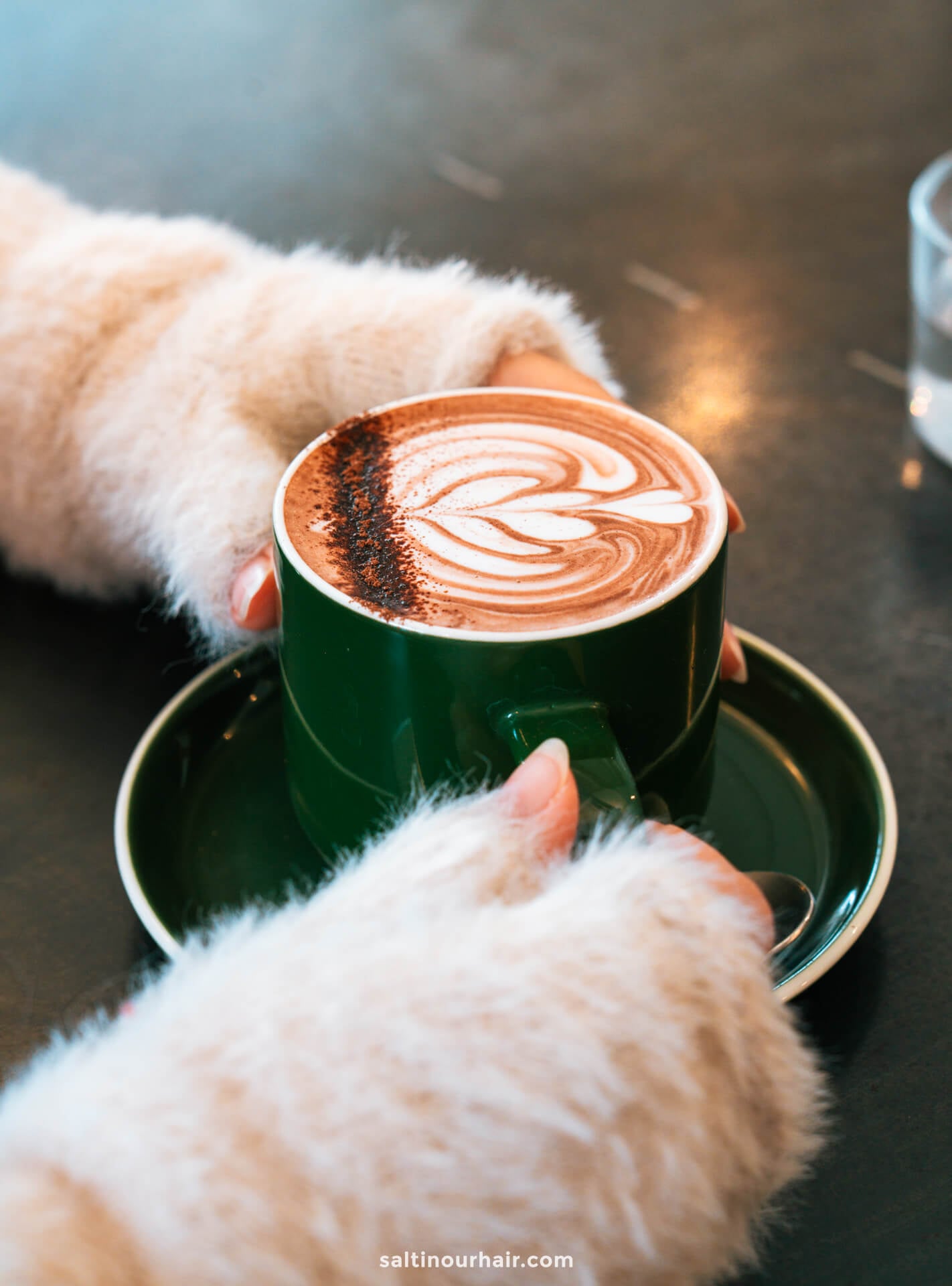 A person wearing a fuzzy light sweater holds a green cup of cappuccino with latte art on a saucer, placed on a dark table in Arrowtown, New Zealand.