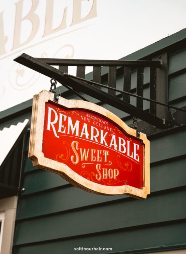 Red and gold hanging sign reads Remarkable Sweet Shop, Arrowtown, New Zealand, mounted on a green and white exterior wall&mdash;a delightful stop among things to do Queenstown.