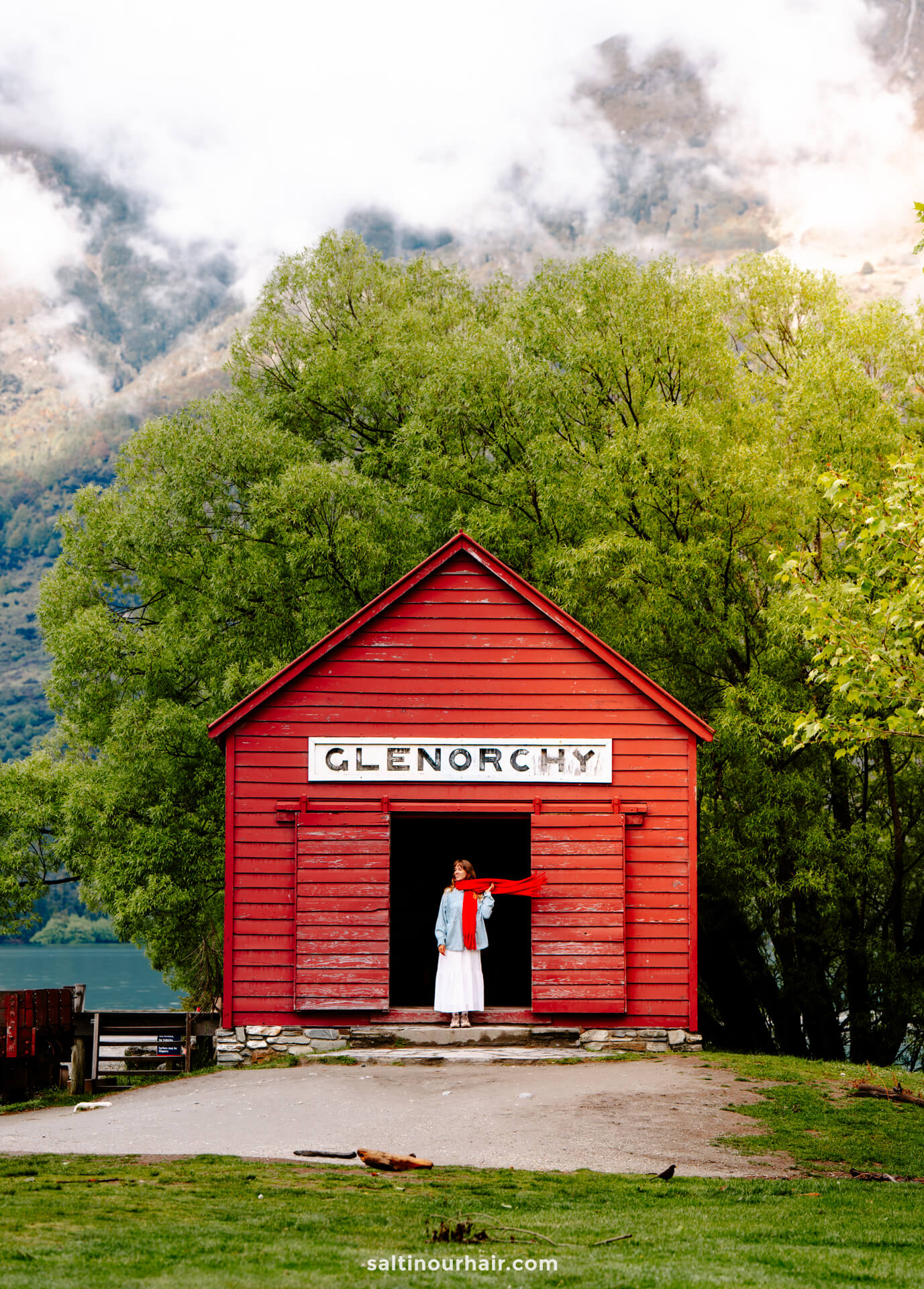 A person stands in the doorway of a red wooden building labeled GLENORCHY&mdash;one of the iconic things to do Queenstown offers&mdash;surrounded by green trees and mountains in the background.
