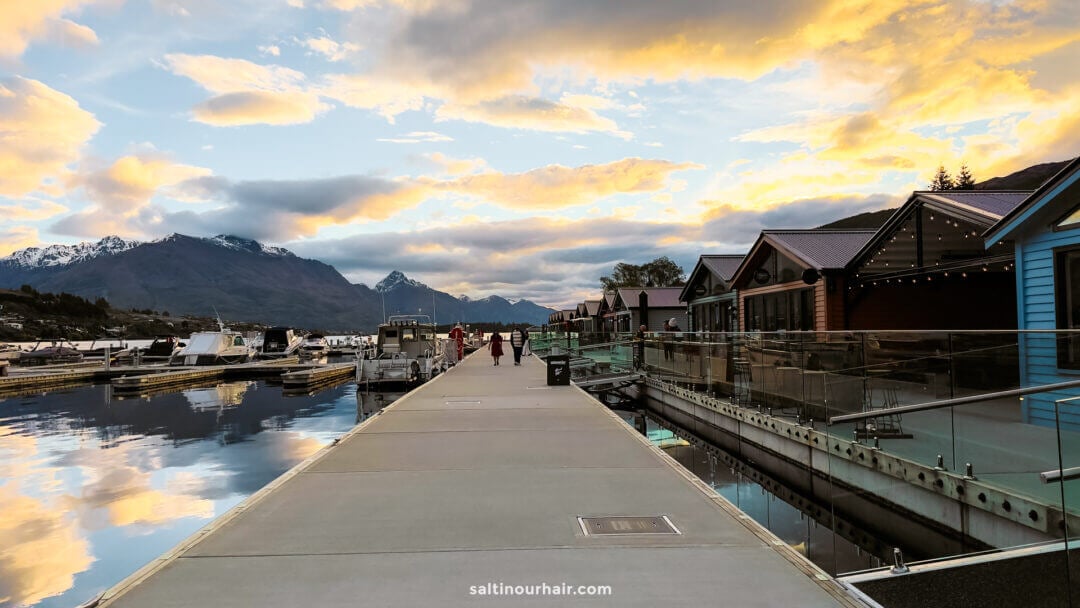 A dock with moored boats on a calm waterfront, lined with modern buildings and set against mountains at sunset&mdash;one of the many picturesque things to do in Queenstown under a cloudy sky.