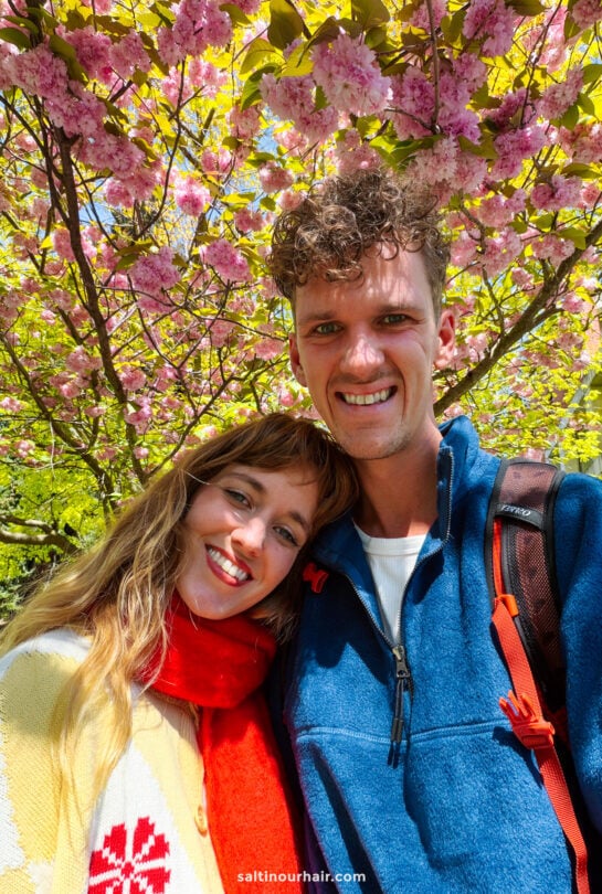A smiling couple stands under blooming pink cherry blossom trees, with sunlight filtering through the branches&mdash;capturing one of the many enchanting things to do in Queenstown.